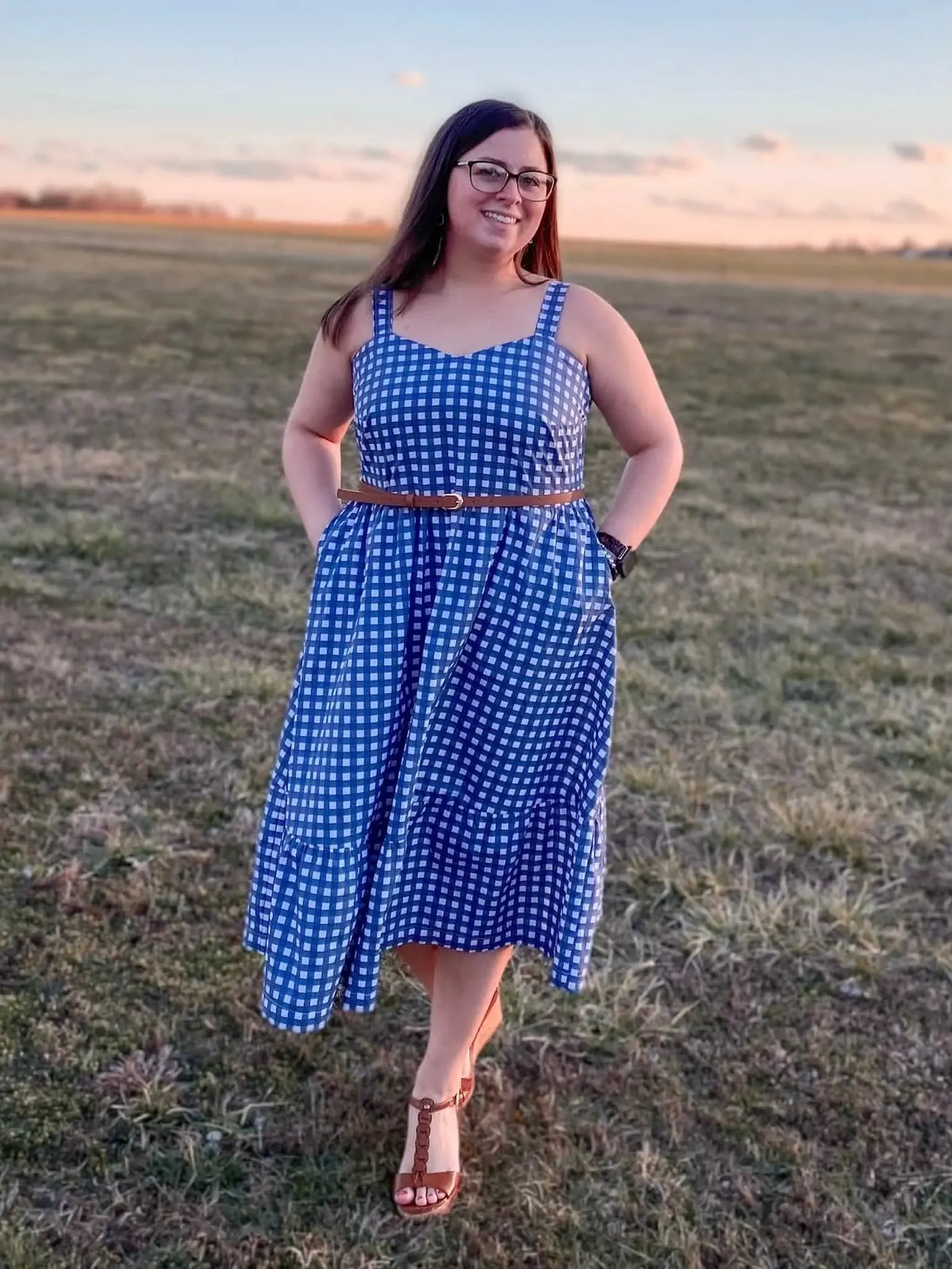 A woman in a navy gingham dress with a brown belt stands in a field.