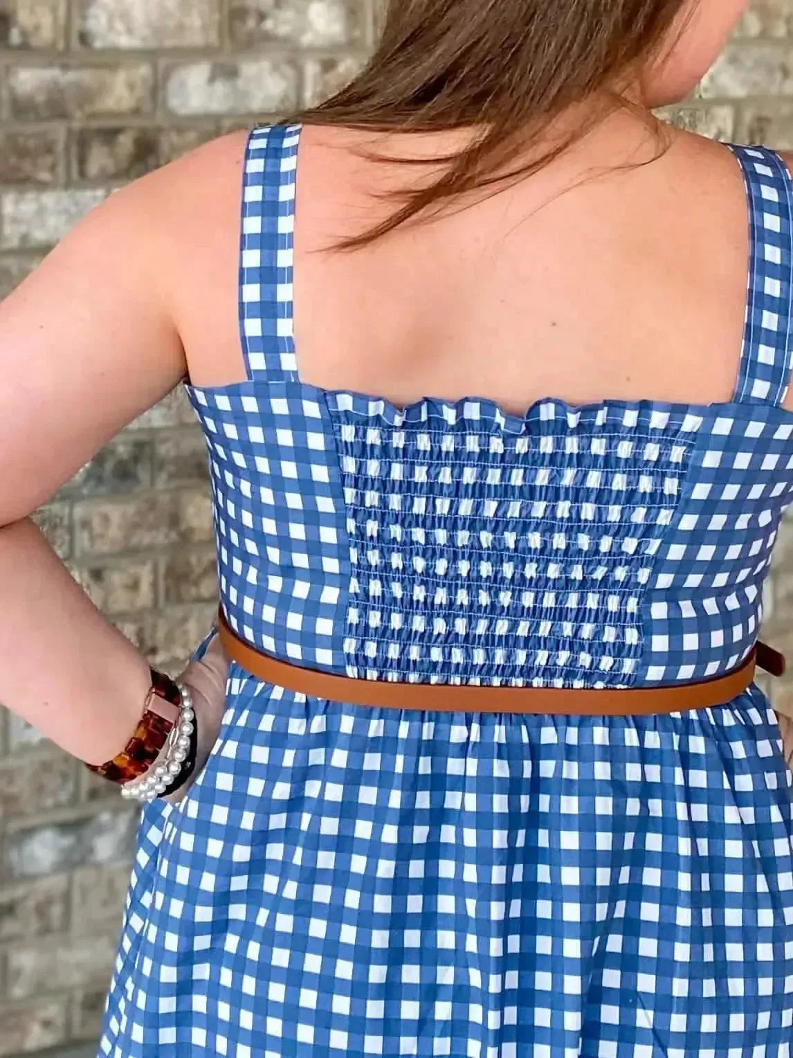 Back view of a woman wearing a navy gingham checkered sundress with brown belt.
