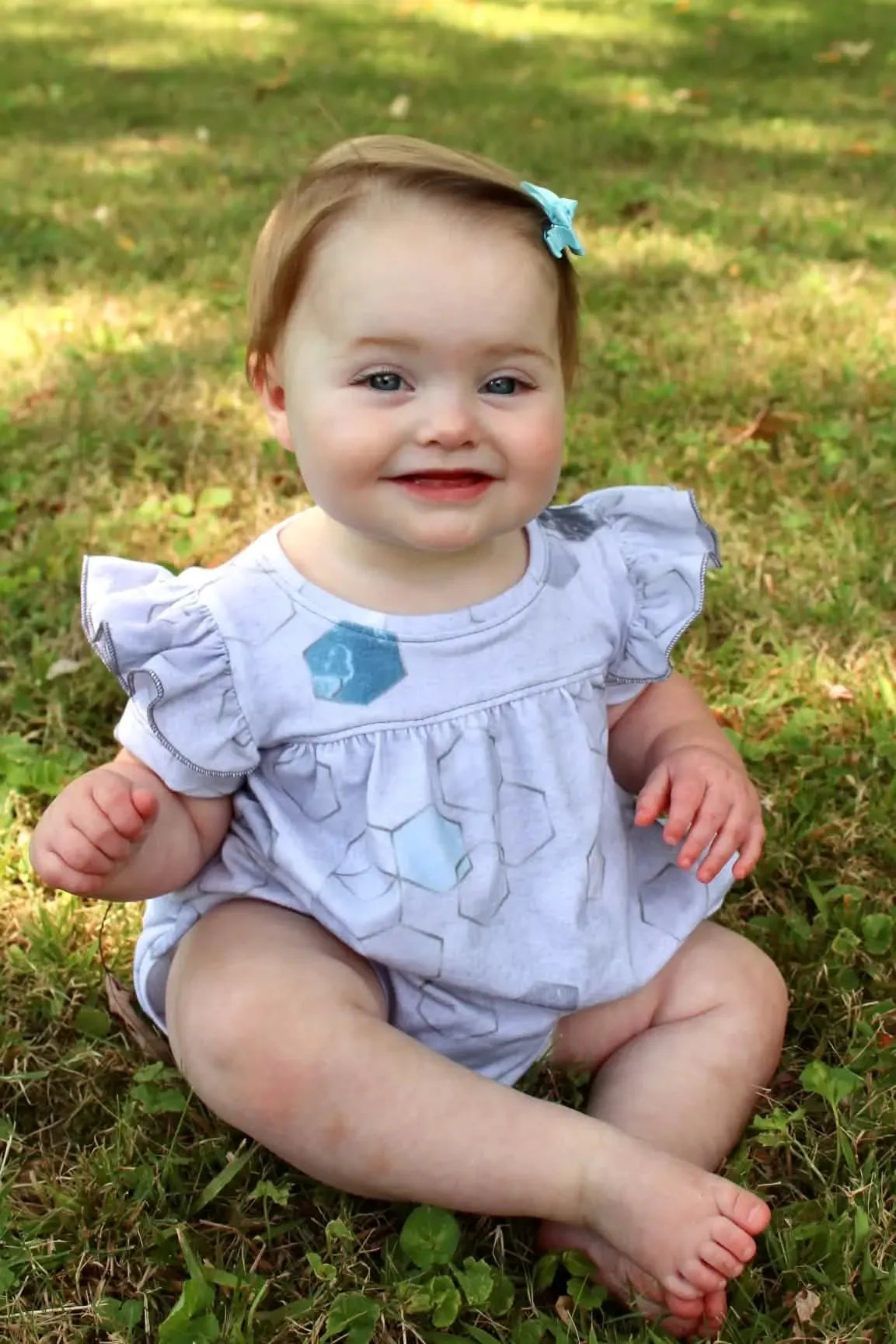 A baby in a silver geo print outfit with a blue bow in her hair sits on grass.