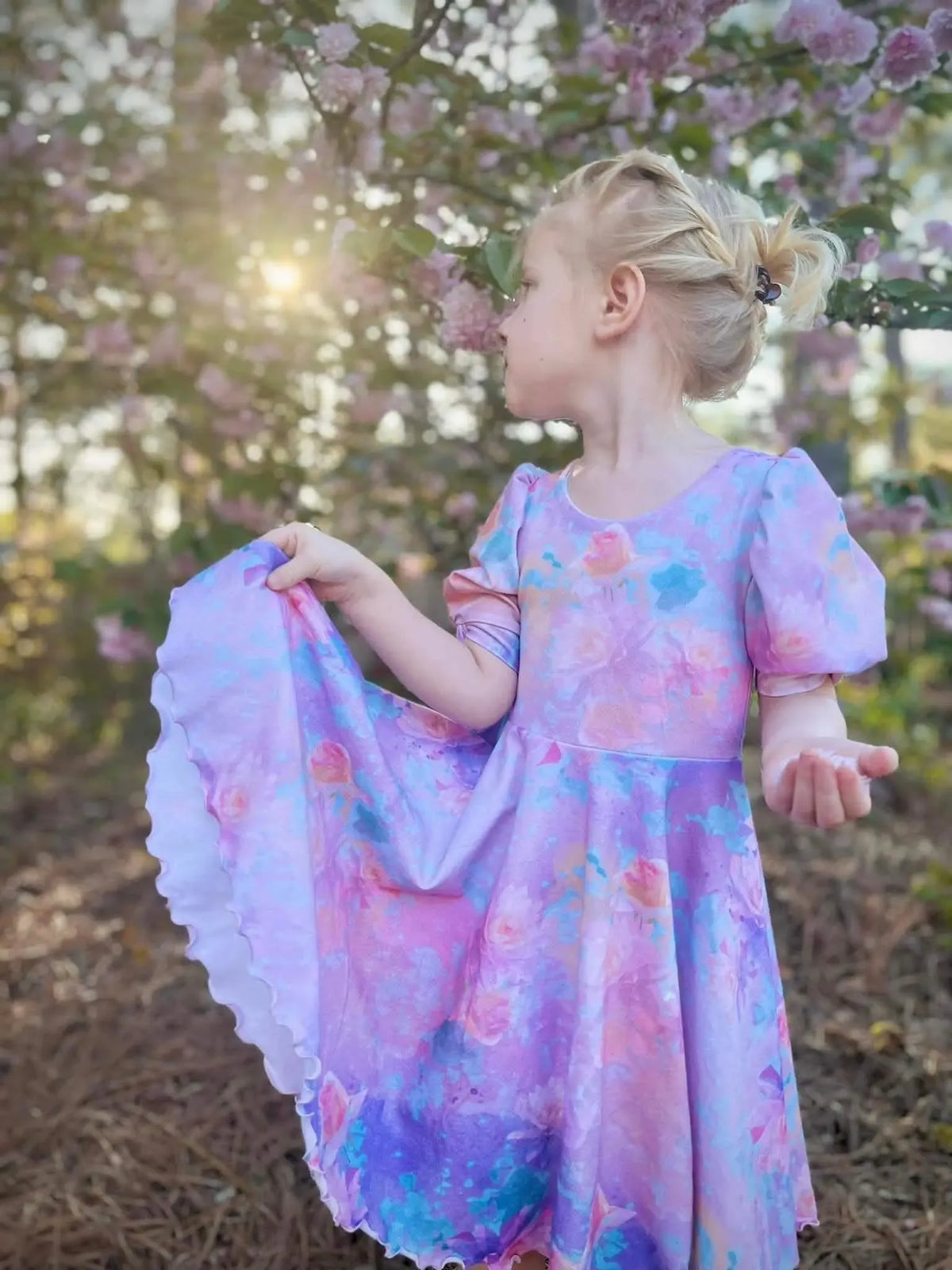 A young girl in a purple floral dress stands among pink flowering trees.