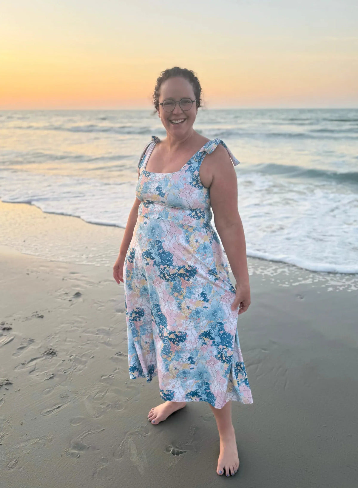 A woman in a floral dress stands on a beach at sunset.