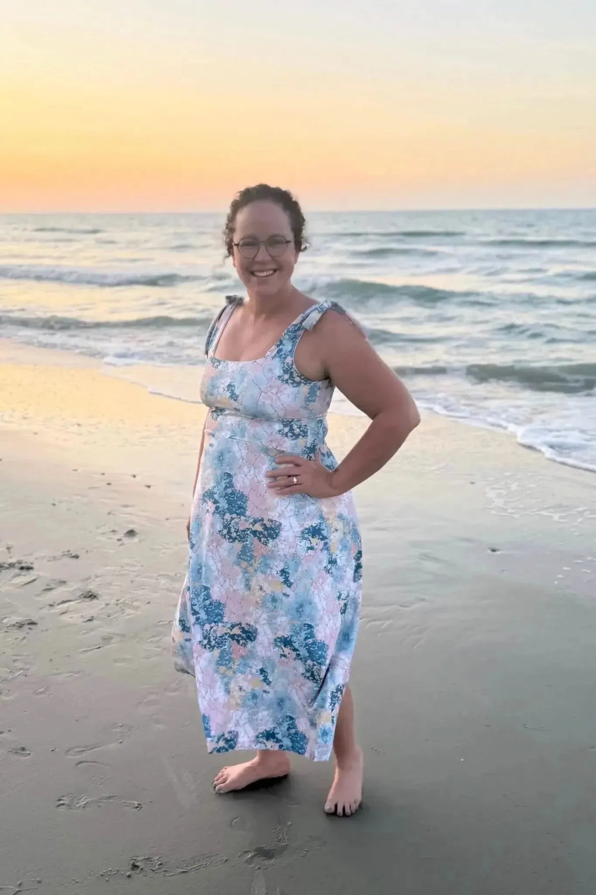 A woman in a floral dress stands on a beach at sunset.