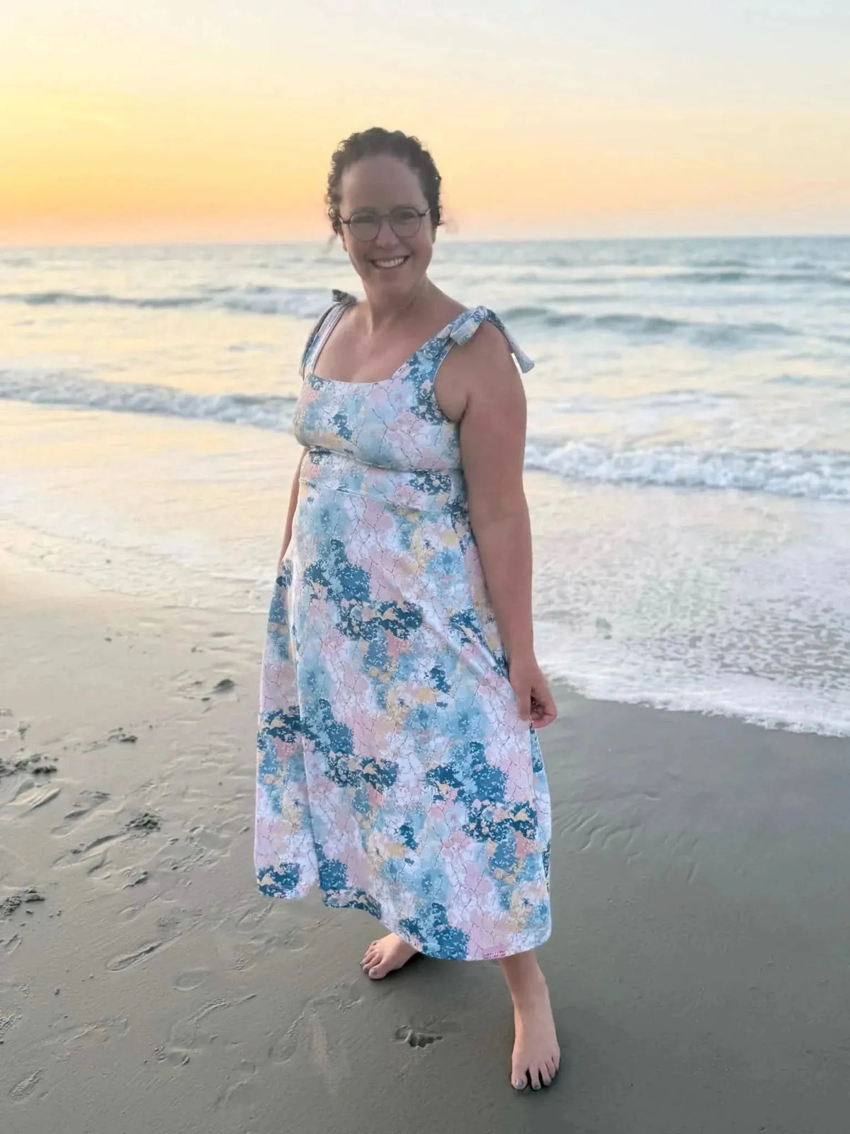 Woman wearing a sleeveless midi dress in abstract blue-pink floral print on beach at sunset.