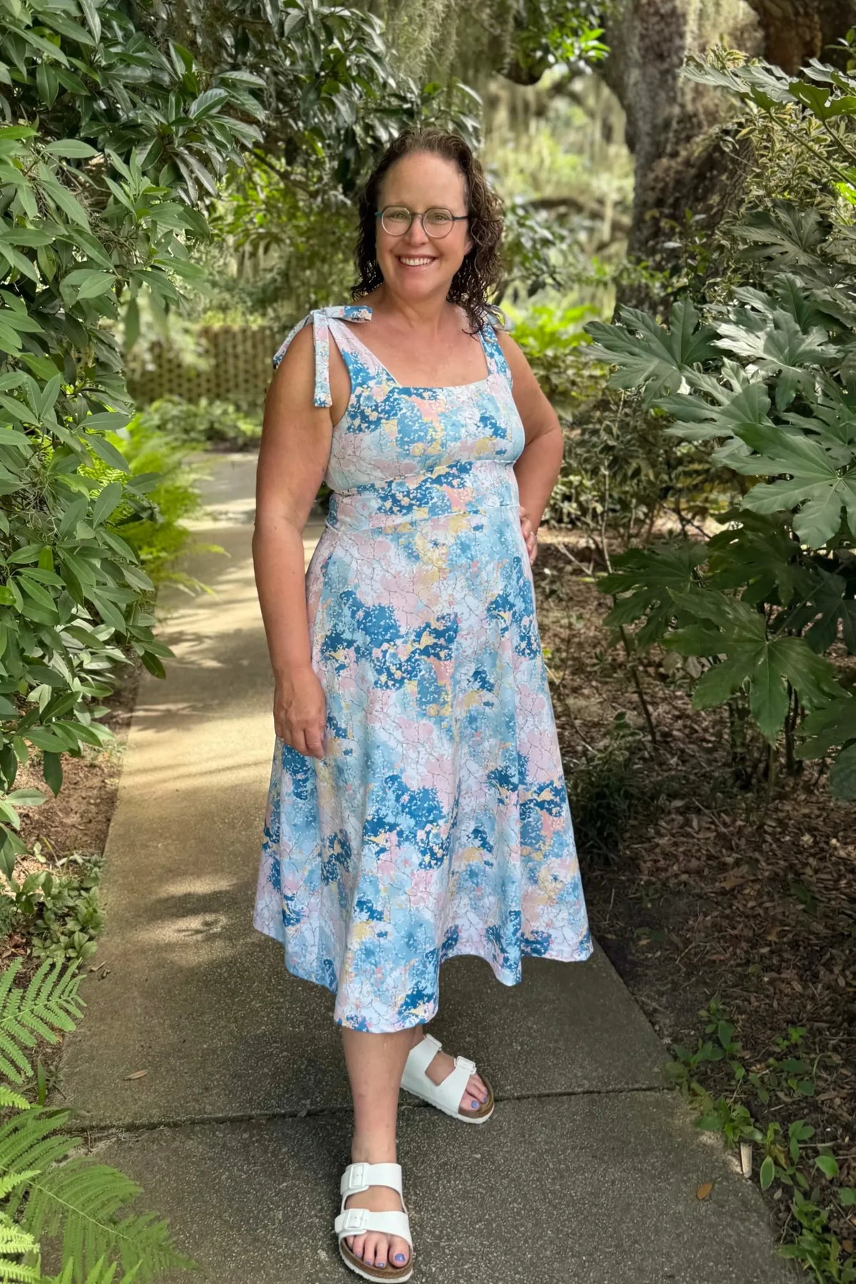 A woman in a blue, pink, and yellow floral dress stands on a path.