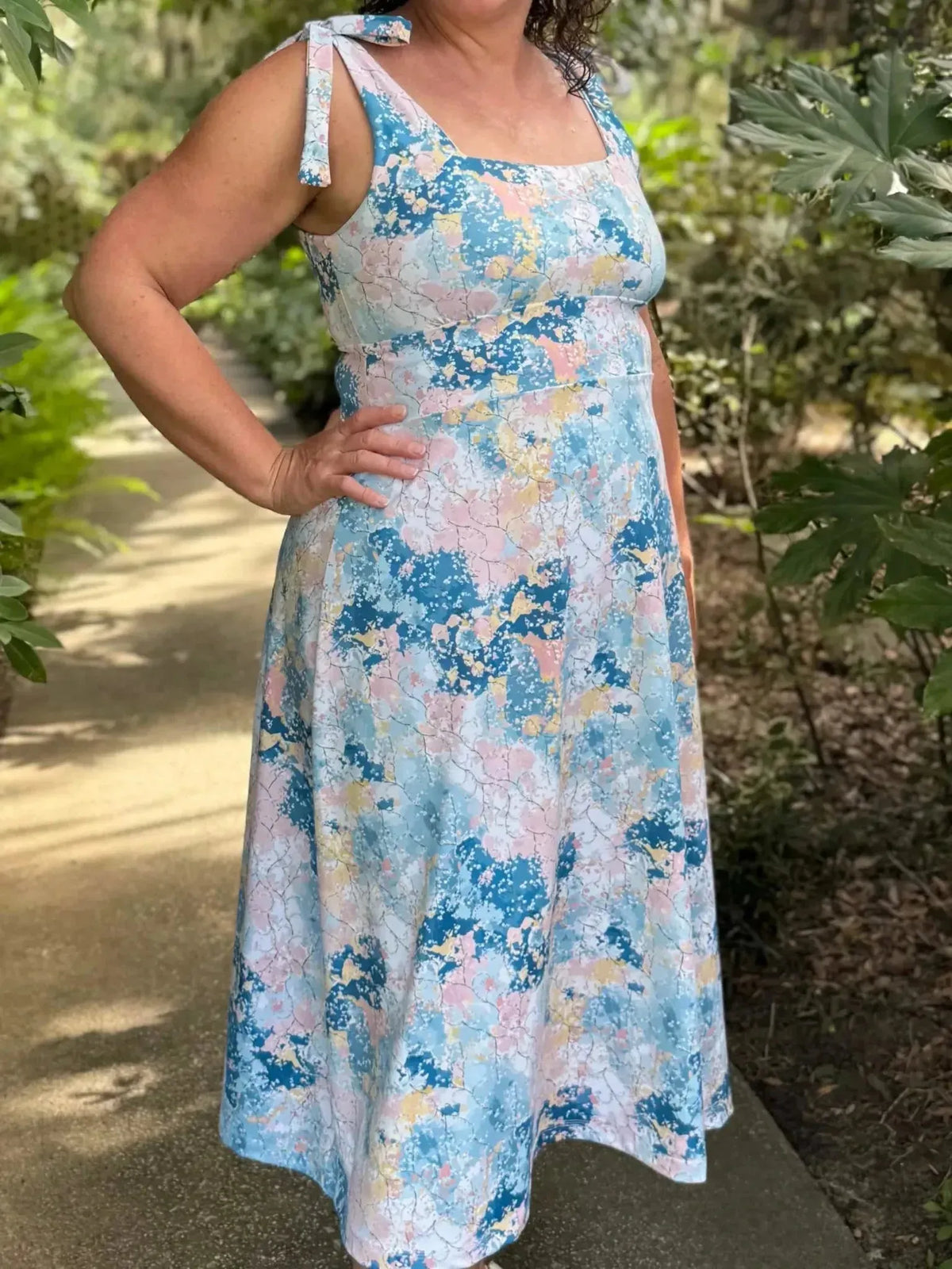Woman wearing a sleeveless blue and pink abstract floral print dress with tie straps in a garden.