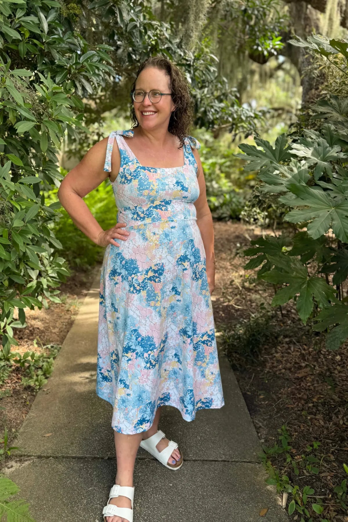 A woman in a colorful floral dress and white sandals stands on a path.