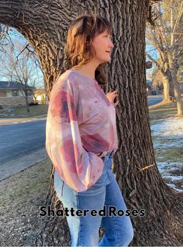 A woman in a pink floral top and jeans leans against a tree.