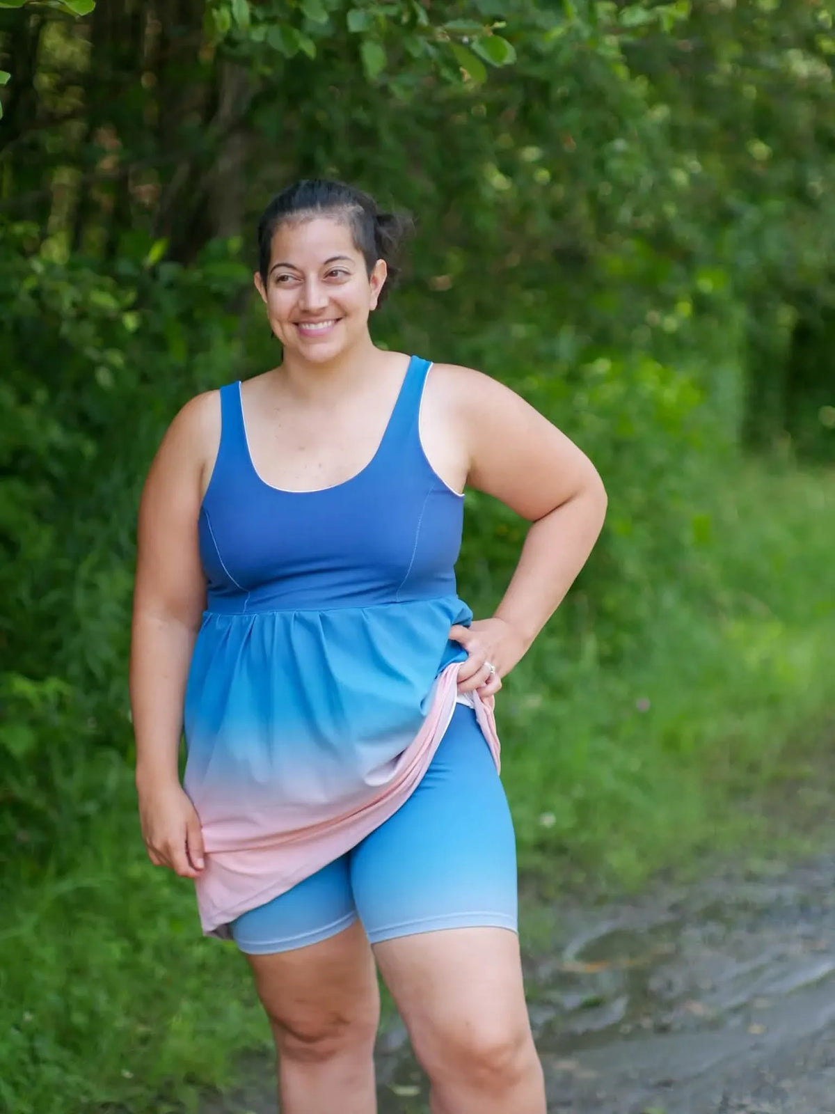 A smiling woman wears a blue and pink ombre tank top and blue shorts outdoors.