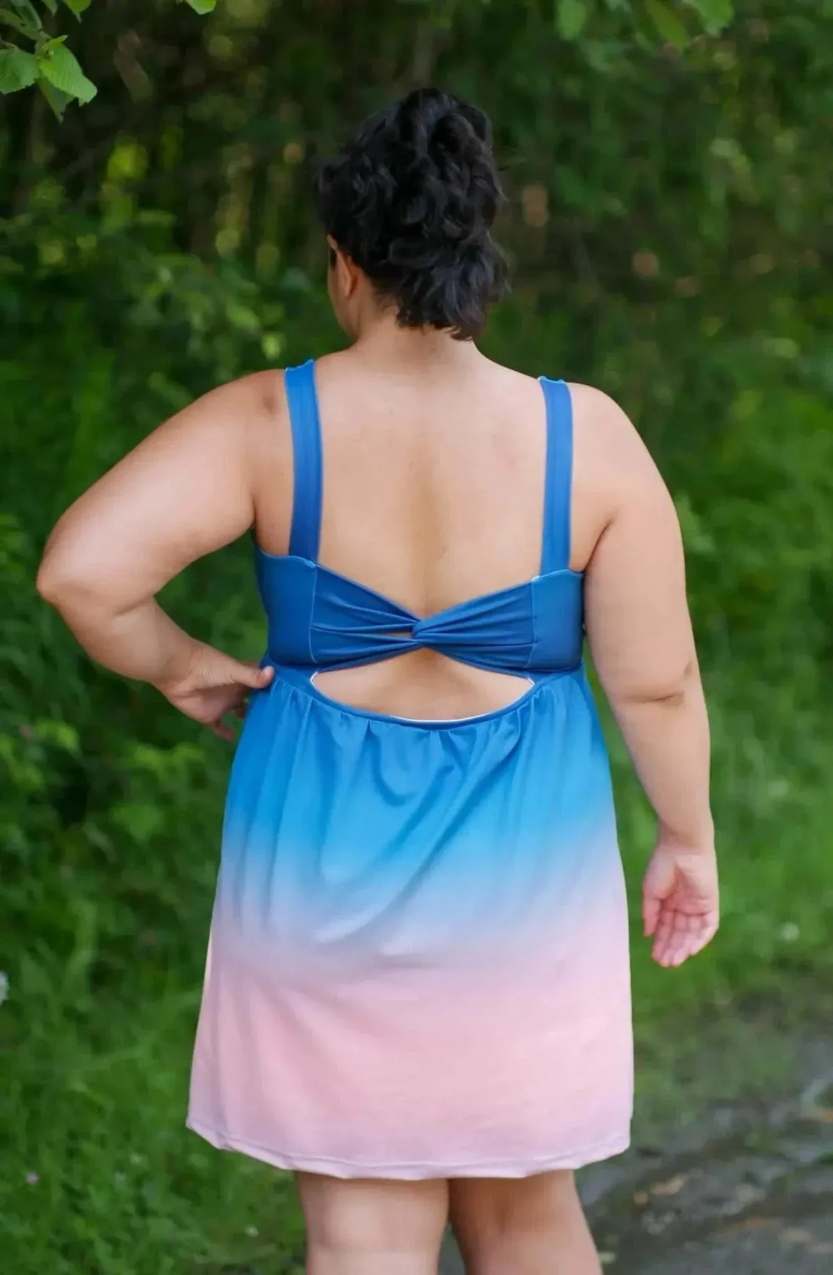 Rear view of a woman wearing a blue-to-pink ombre gradient dress with a bow back detail in a green outdoor setting.