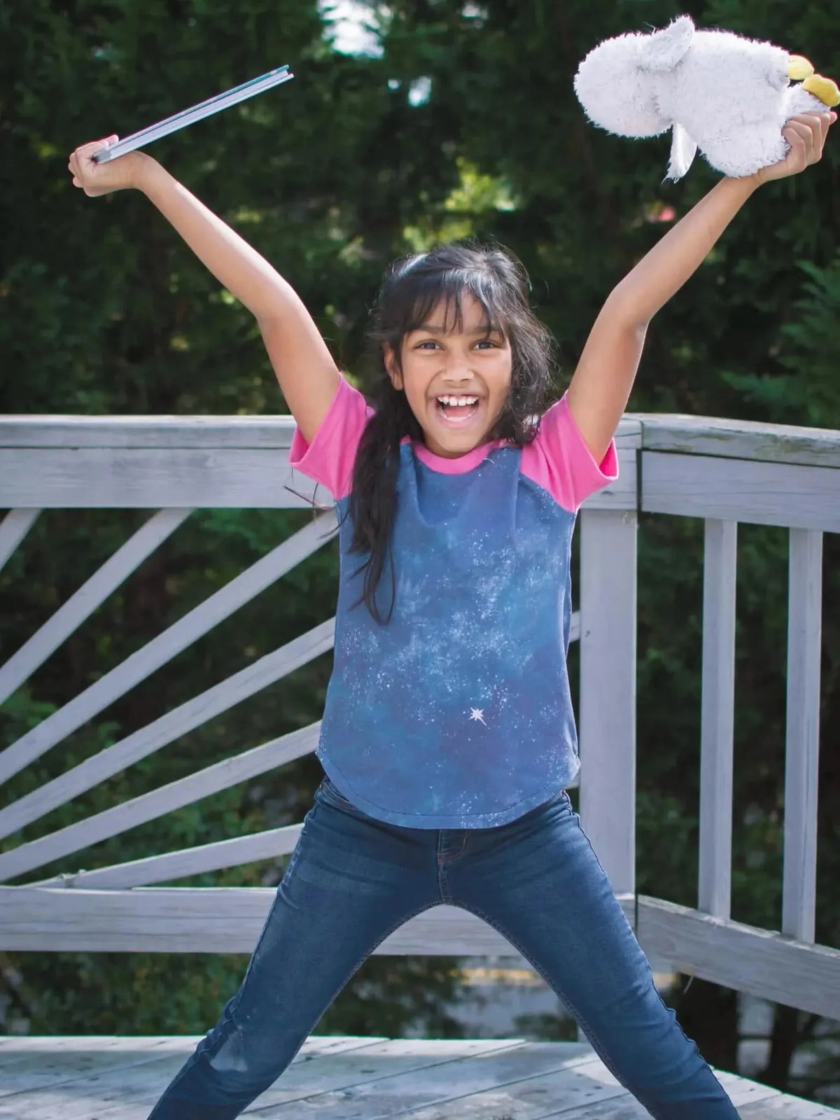 A girl in a blue and pink shirt with a celestial pattern holds a book and a stuffed animal.