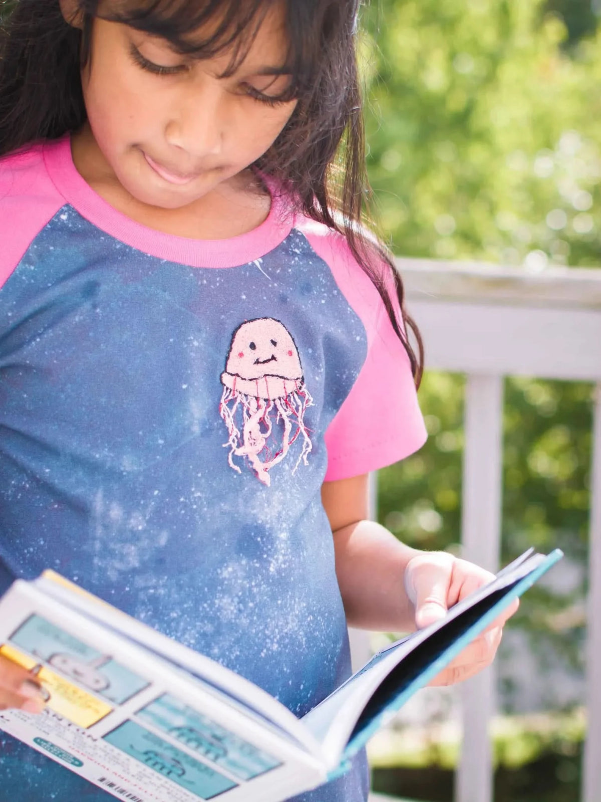 A young girl in a blue and pink shirt with a jellyfish patch reads a book outdoors.