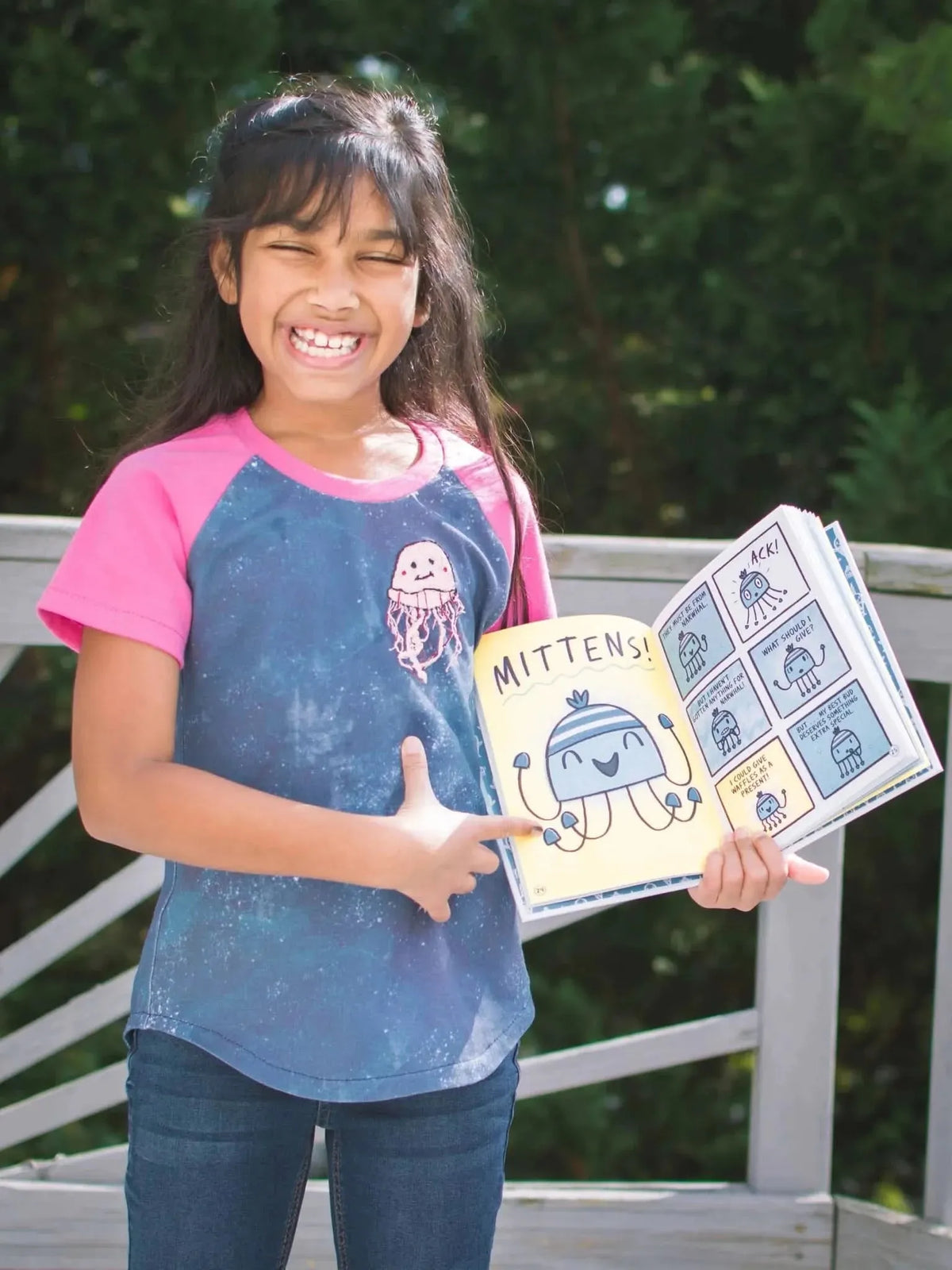 Girl wearing blue t-shirt with pink sleeves and jellyfish print, holding open Mitten book.