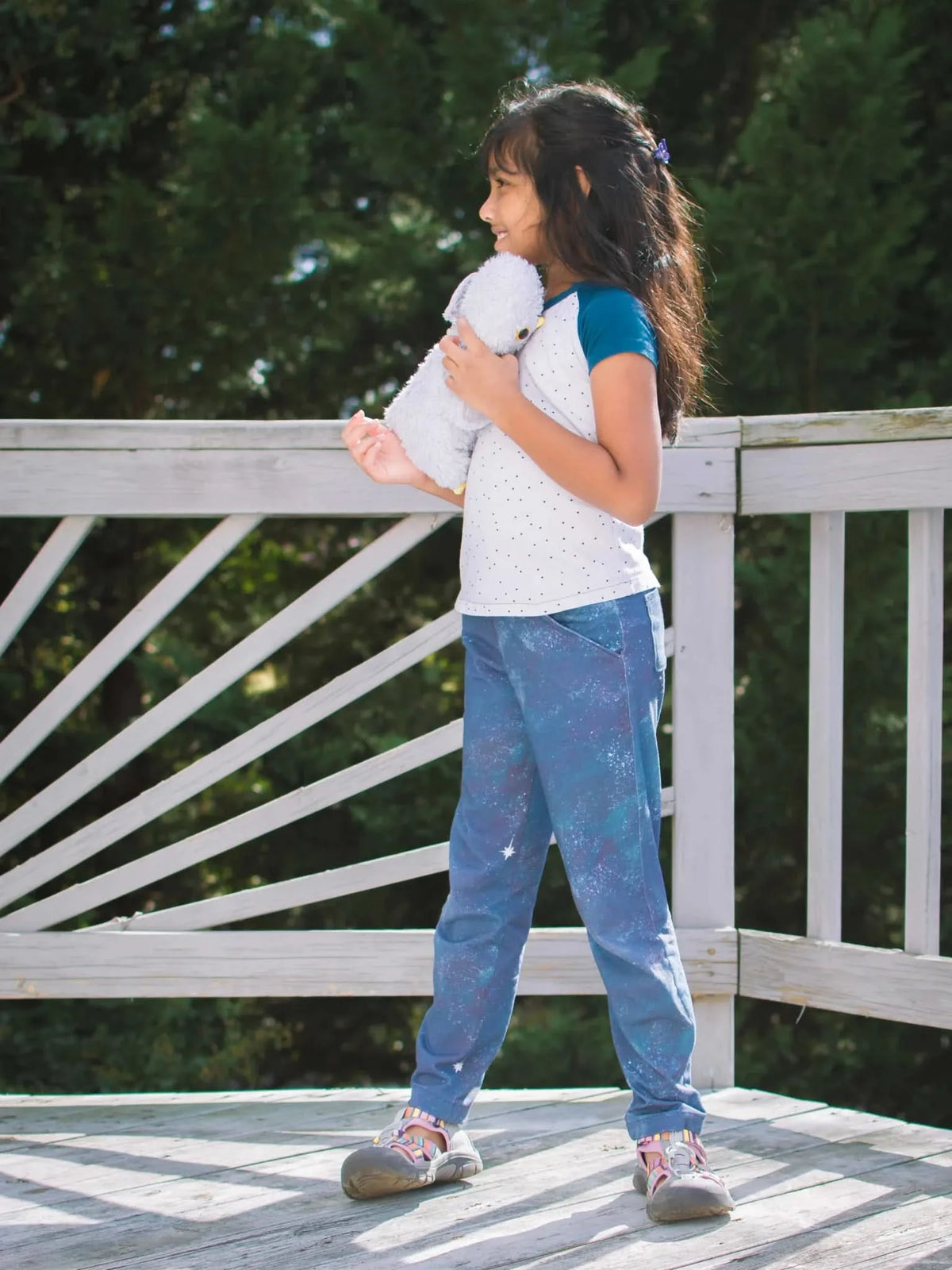 A young girl in a white and blue shirt and blue pants holds a gray stuffed animal.
