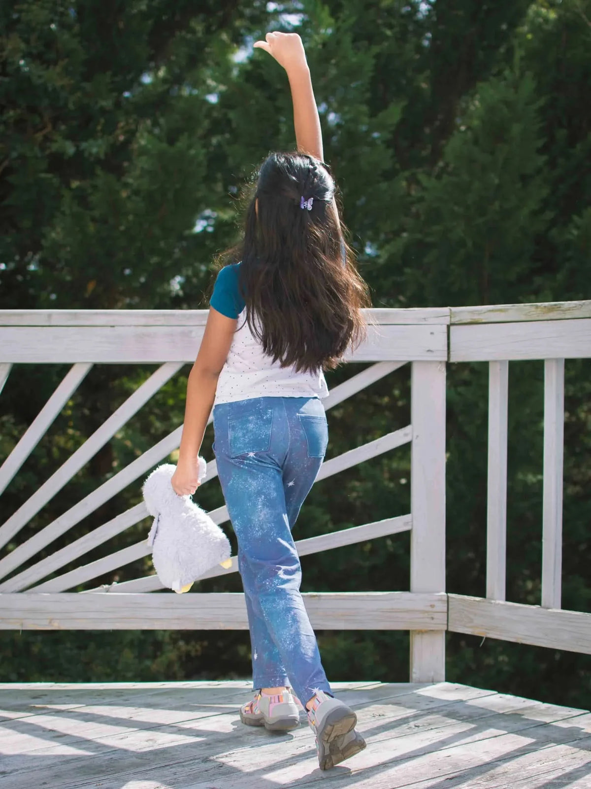 A young girl in blue pants with white star patterns raises her arm on a wooden deck.