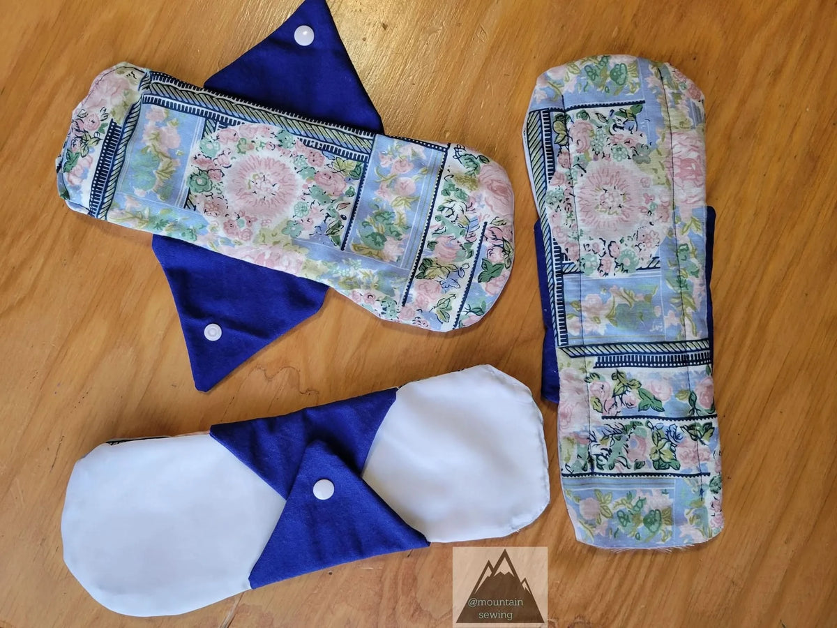 Three reusable menstrual pads with floral patterns and blue wings are displayed on a wooden surface.