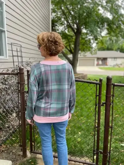 A person from behind wears a dark Christmas plaid shirt with pink trim and blue jeans.