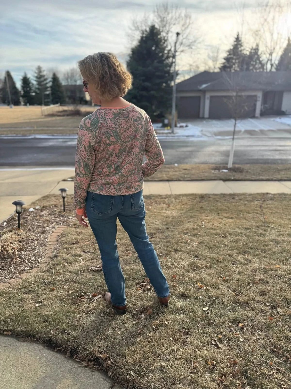 A woman from behind wears a long-sleeved paisley top and blue jeans outdoors.