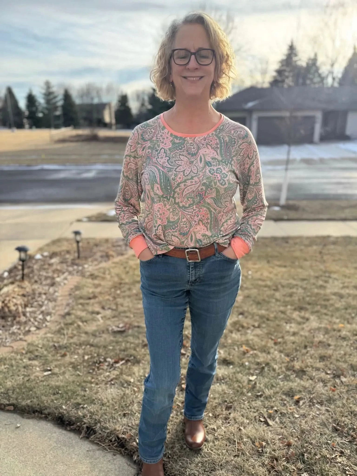 A woman wears a long-sleeved paisley top with jeans and a brown belt.