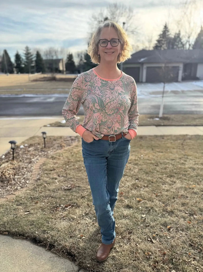 A woman wears a long-sleeved paisley top with orange cuffs and collar, blue jeans, and brown boots.