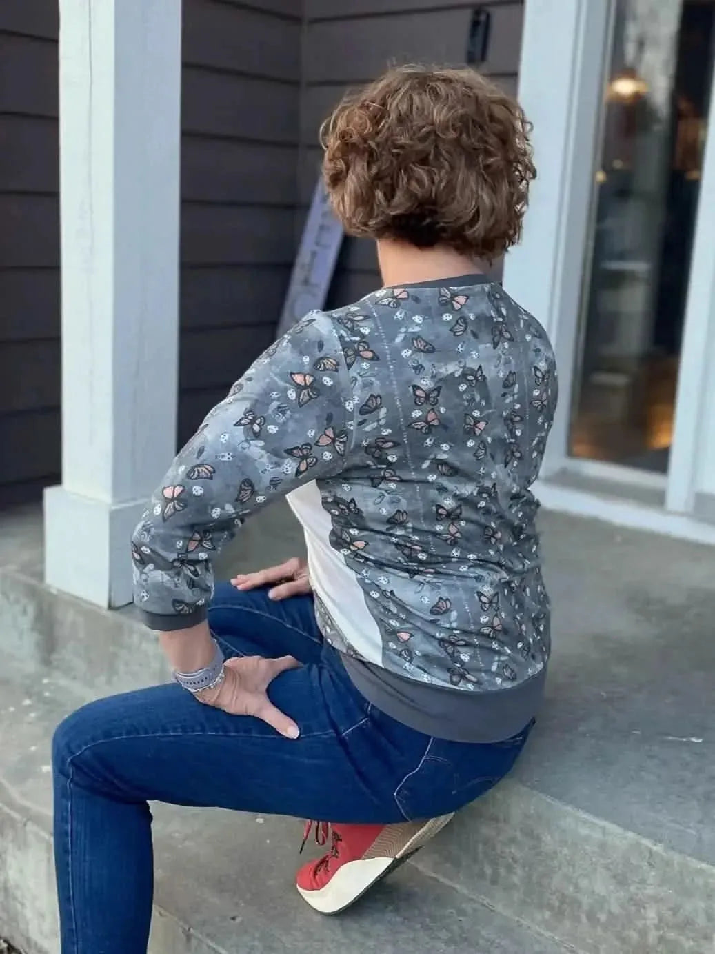 Rear view of woman wearing gray monarch butterfly print jacket and jeans, seated on porch steps.