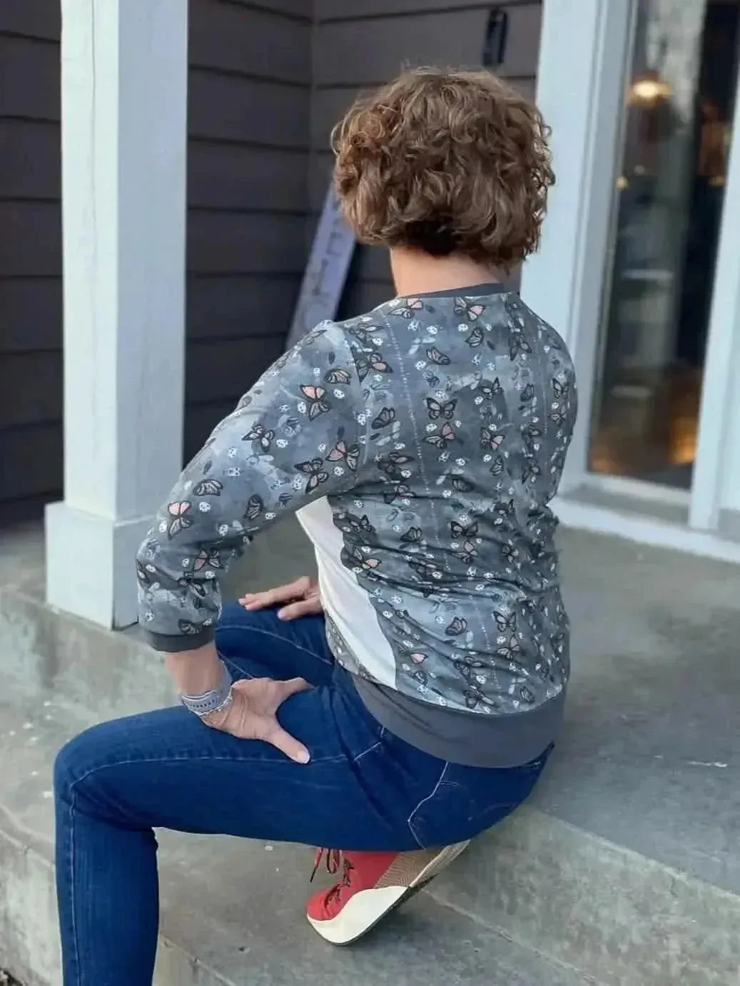 Rear view of woman wearing gray monarch butterfly print jacket and jeans, seated on porch steps.