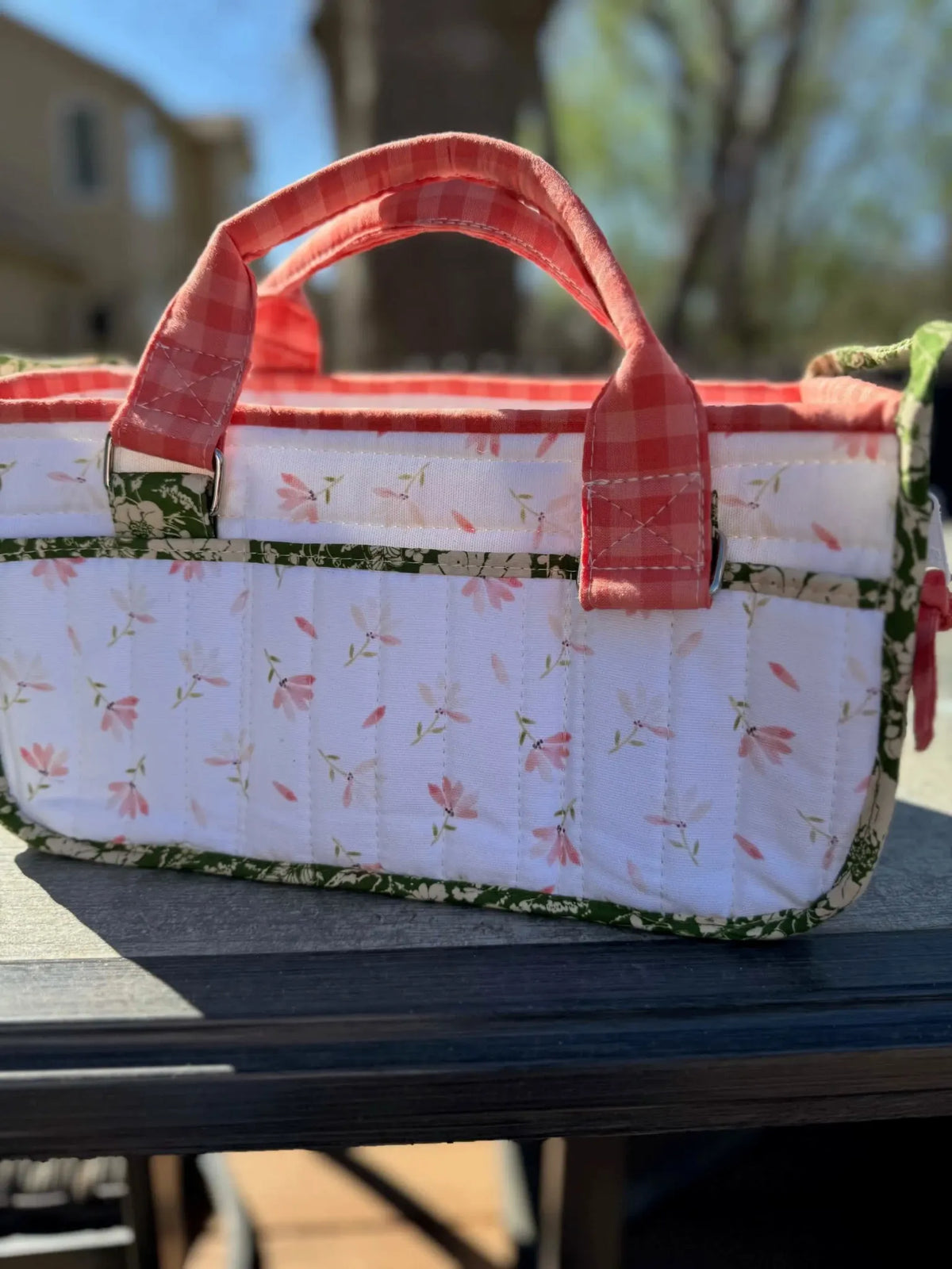 A quilted fabric basket with pink flowers and green trim, featuring a red gingham handle.