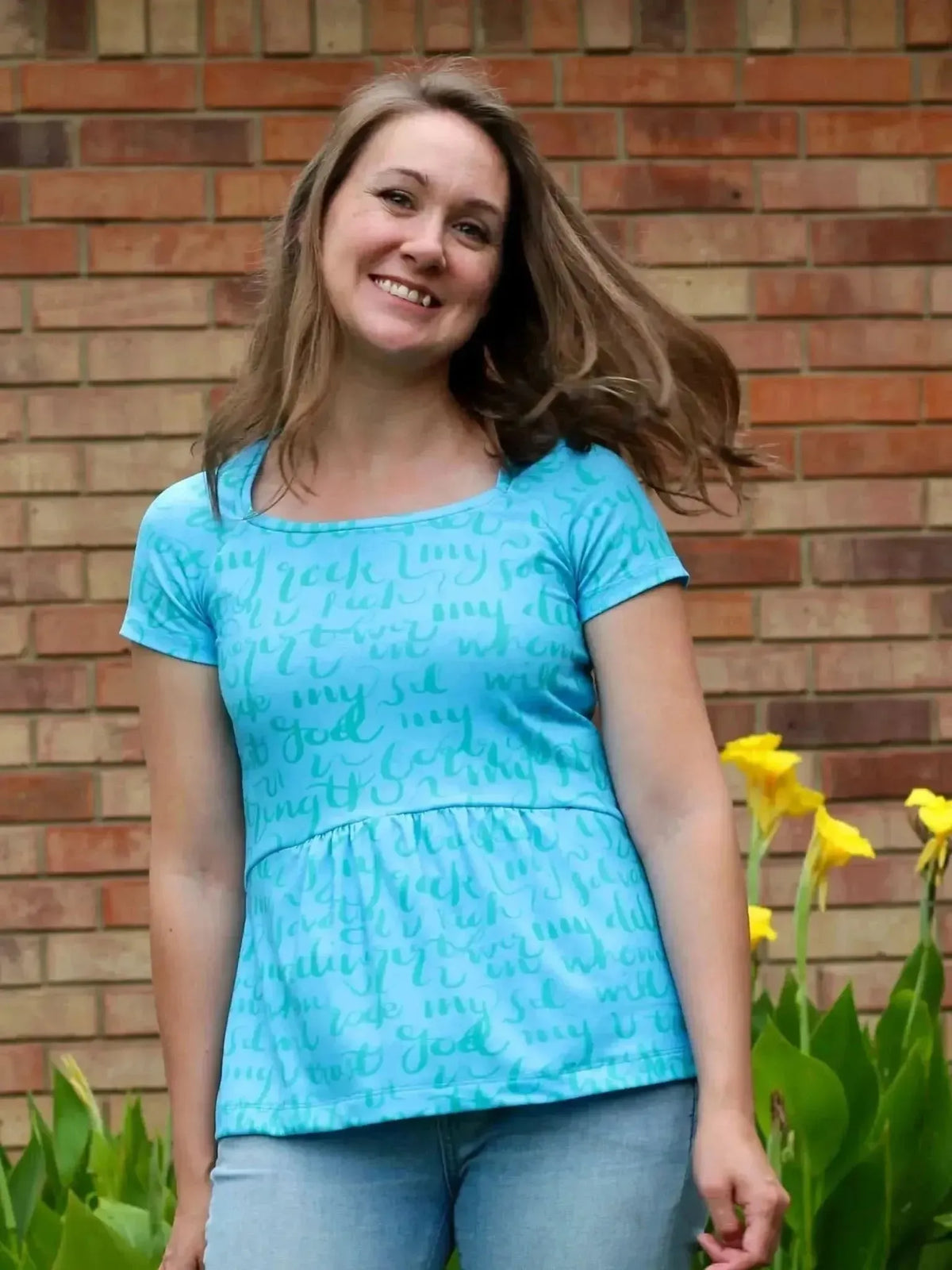 Woman wearing a teal scoop-neck short-sleeve top with "The Lord is My Rock" print against a brick wall.