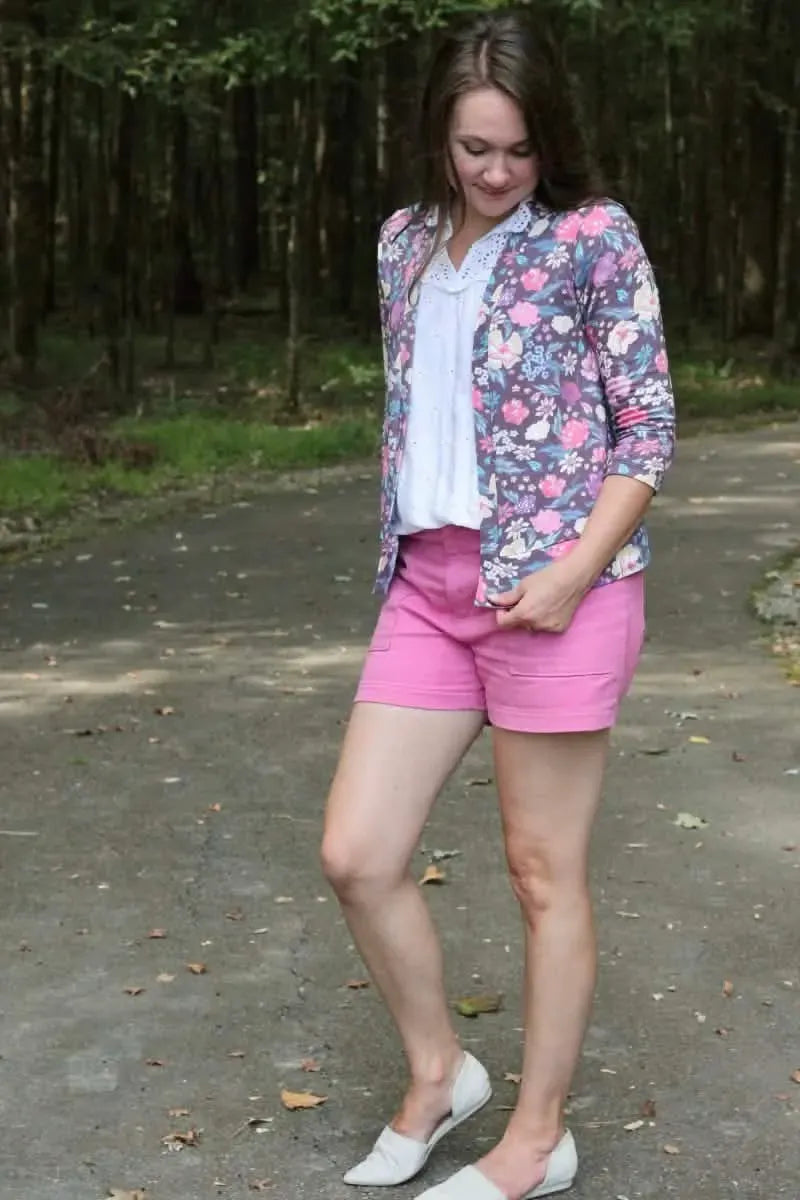 A woman wears a floral print top, pink shorts, and white flats on a path.