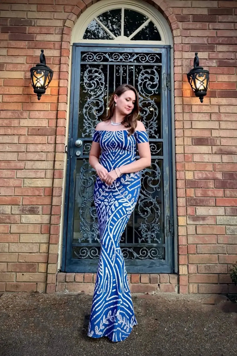 A woman in a blue and white patterned off-the-shoulder dress stands in front of a door.