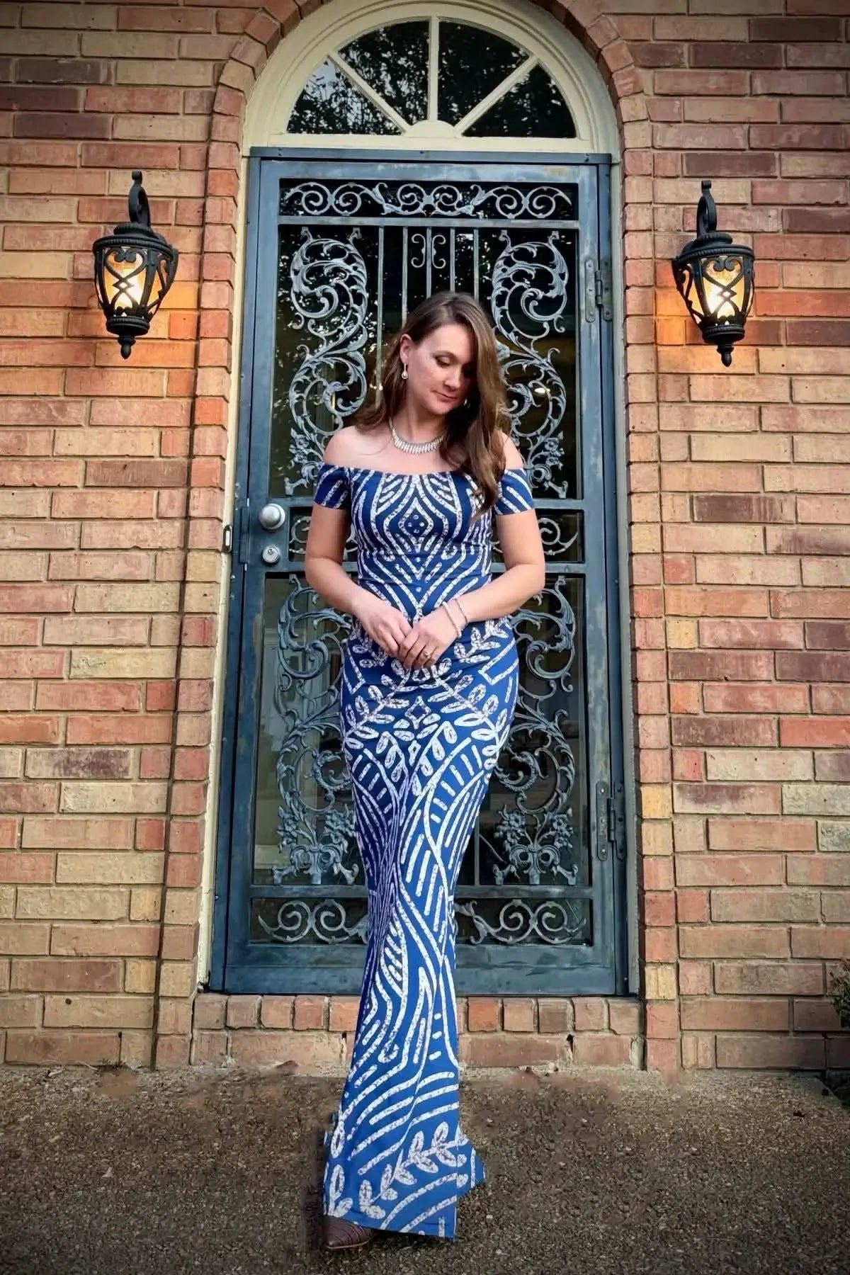 A woman in a blue and white patterned off-the-shoulder dress stands in front of a door.