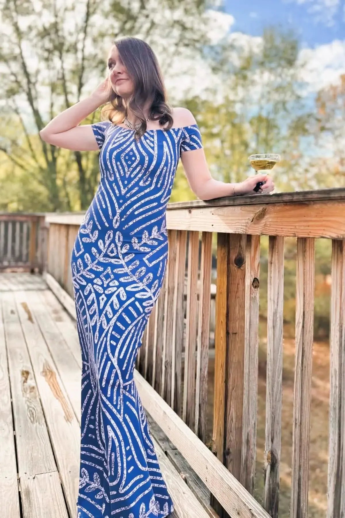A woman in a blue and white patterned dress stands on a wooden deck holding a drink.