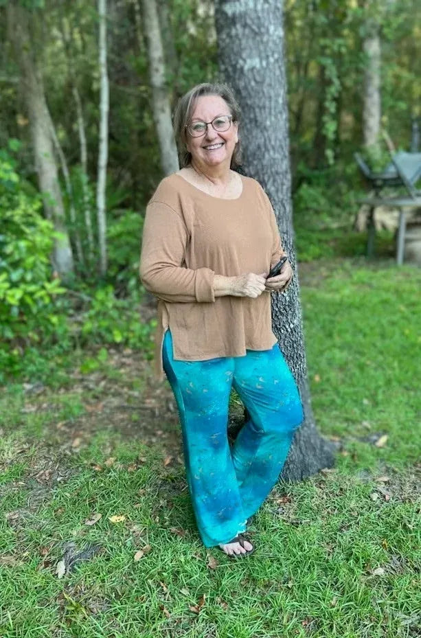A woman in a brown top and dark teal patterned pants stands by a tree.