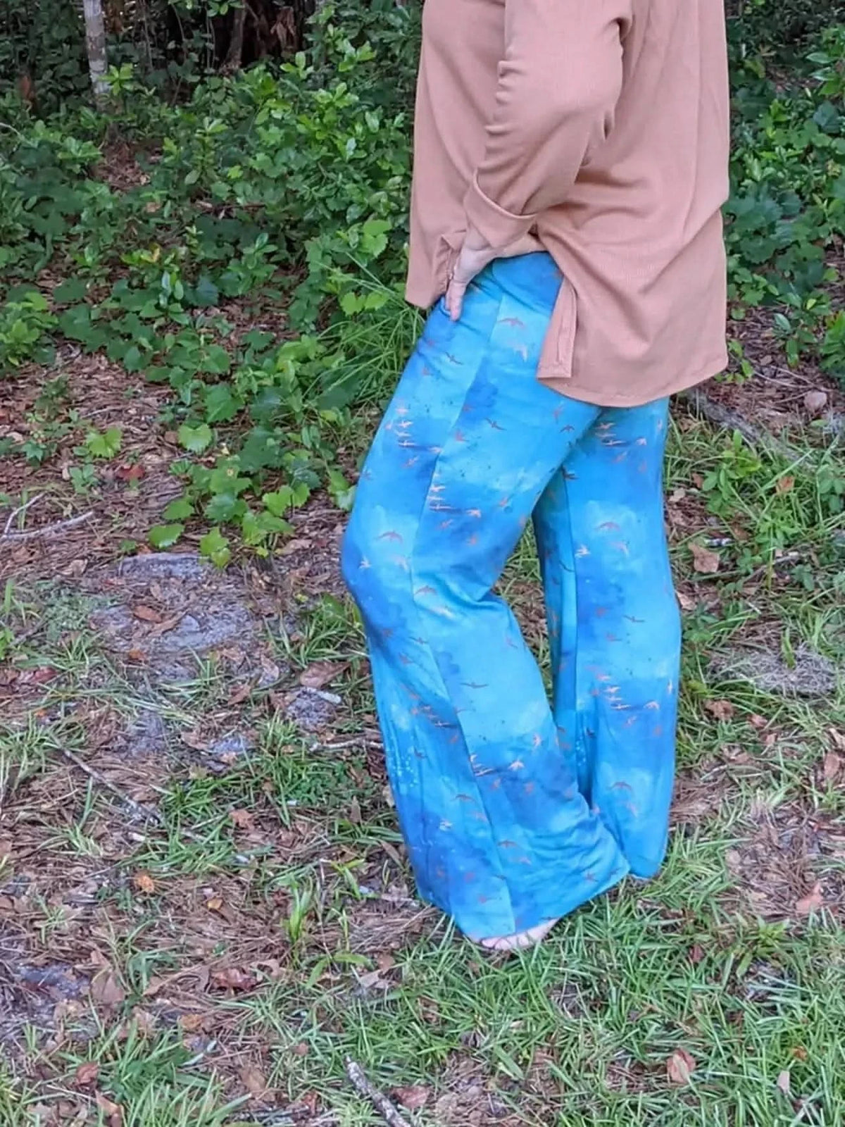 A person wears blue pants with a bird pattern and a tan shirt in a wooded area.