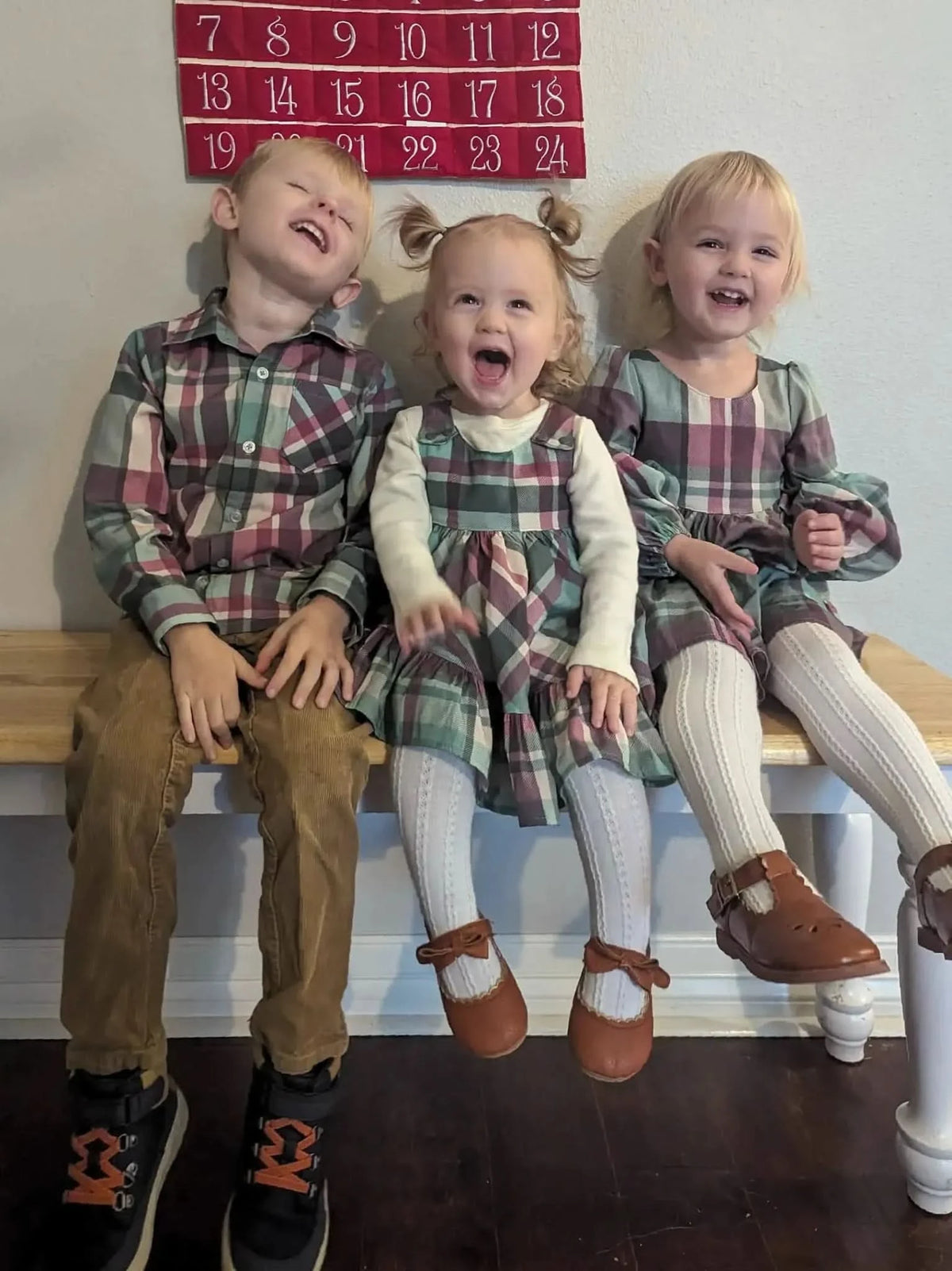 Three children in matching light Christmas plaid outfits sit on a wooden bench.