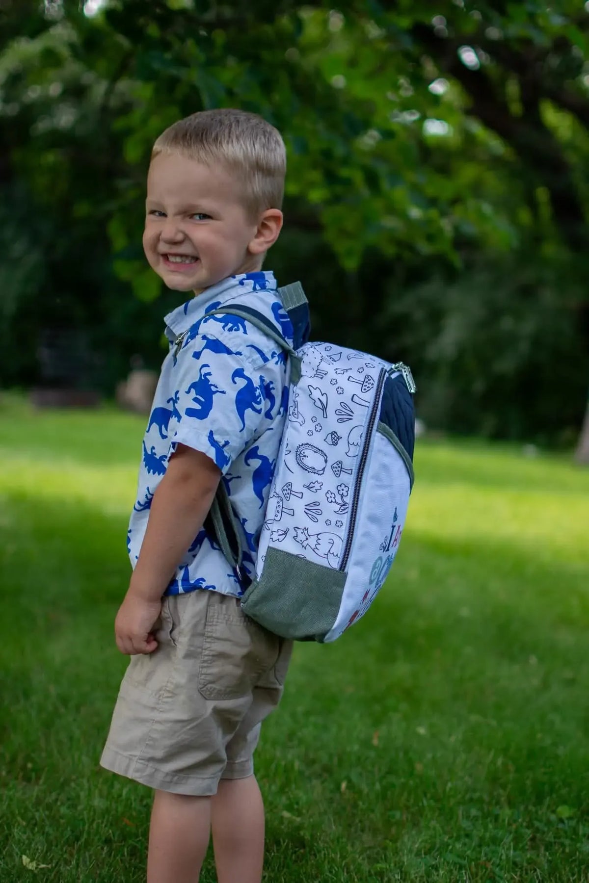 A young boy wears a backpack with a white and green woodland creature pattern.