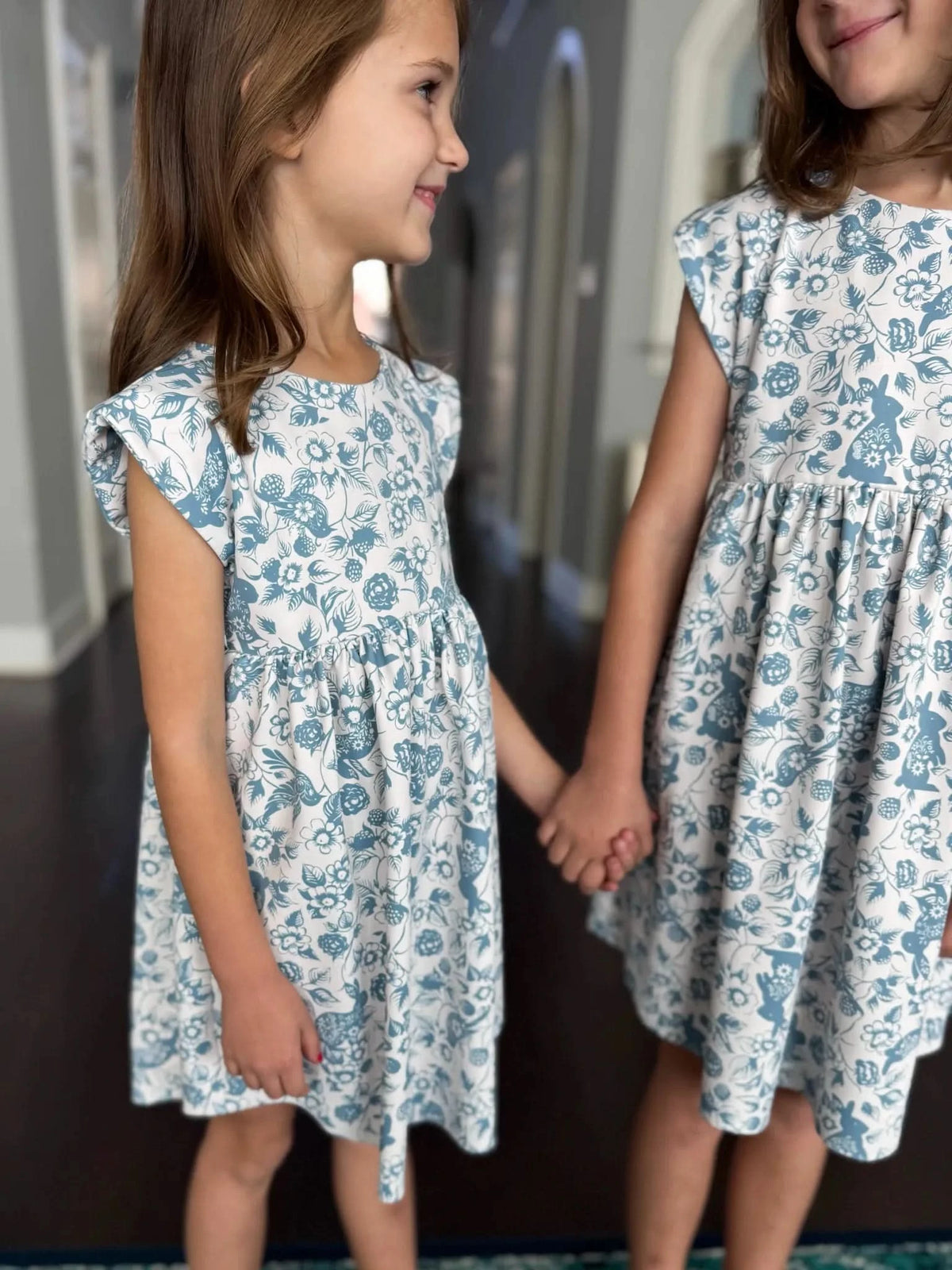 Two girls in blue and white bunny and floral print dresses hold hands.