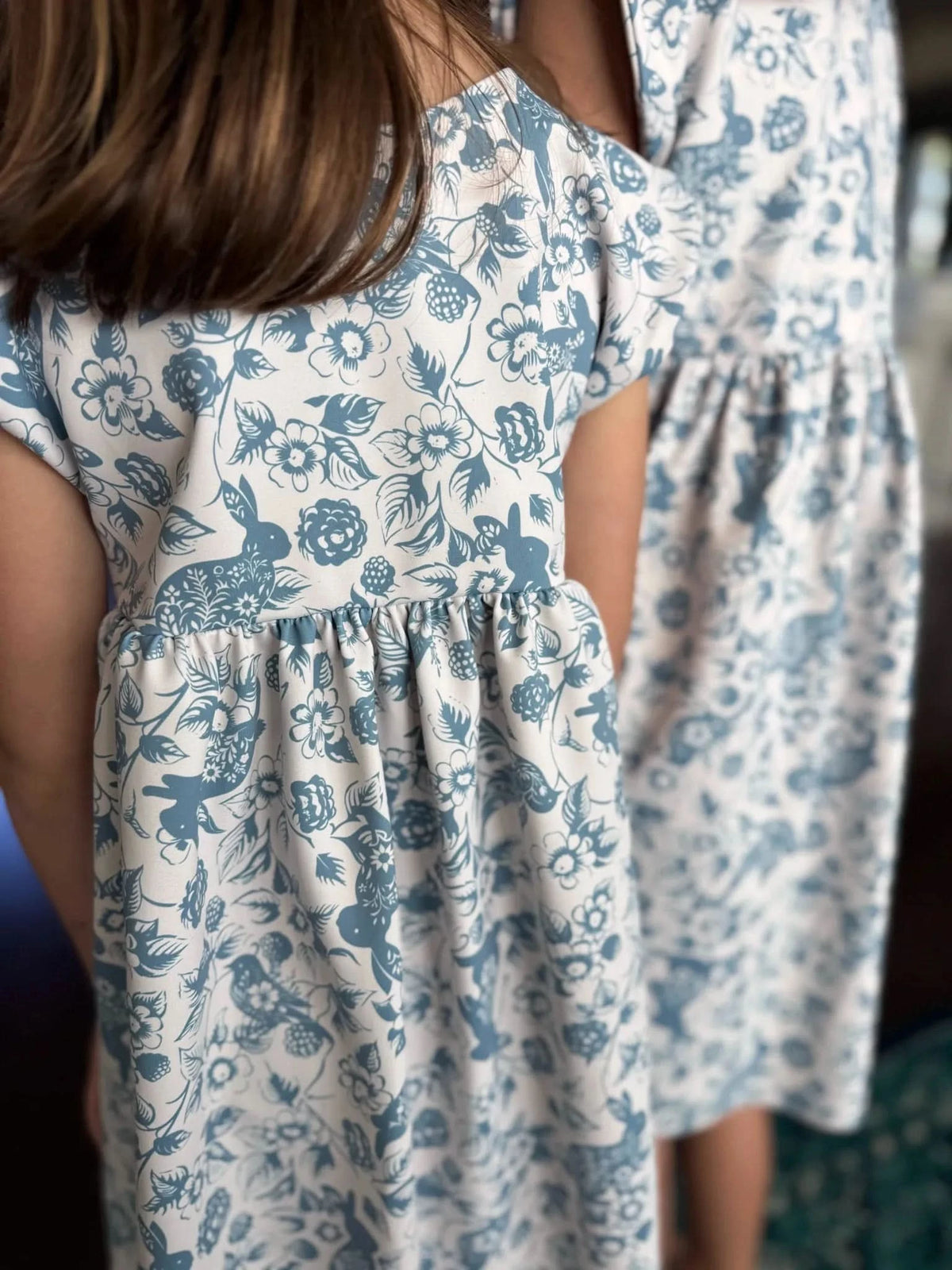 Two people wear dresses with a blue bunny and floral pattern on a white background.