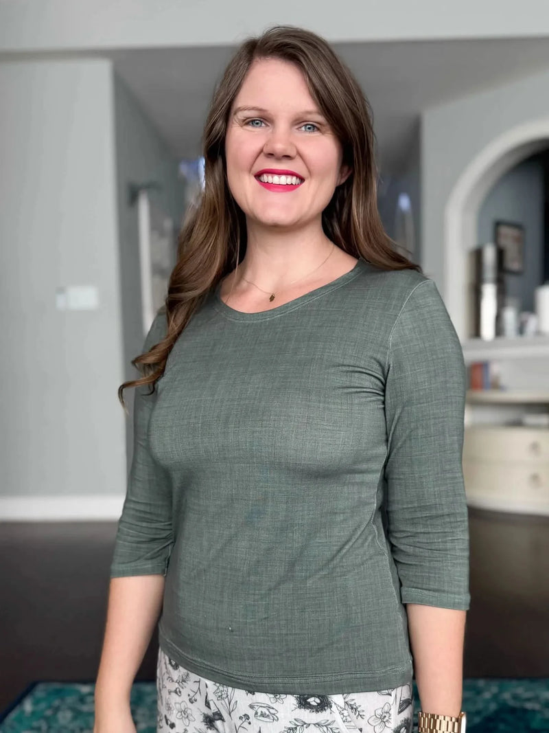 A woman smiles, wearing an olive green shirt and patterned pants.