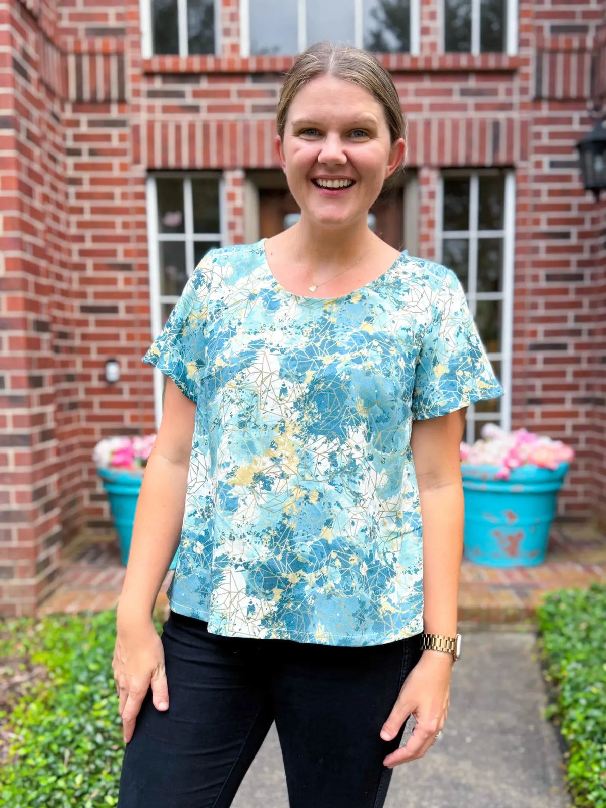 A woman smiles wearing a blue, white, and gold patterned top and black pants.