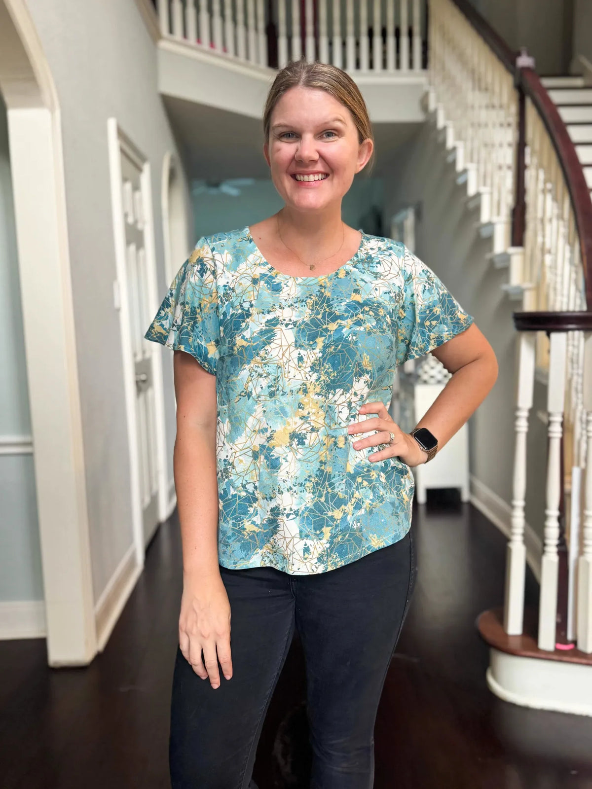A woman smiles wearing a blue, white, and gold patterned top and black pants.
