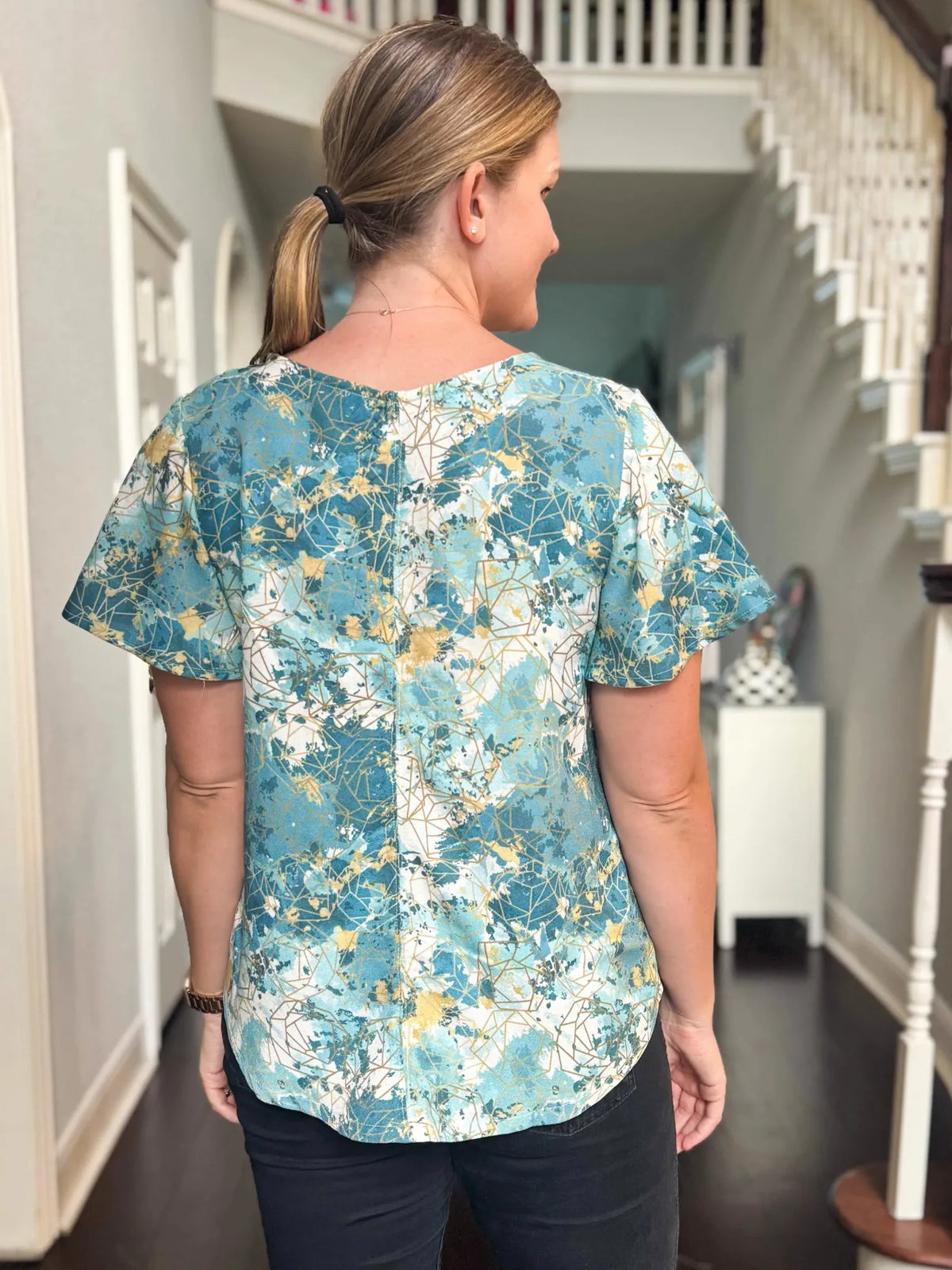 A woman from behind wears a blue, white, and gold patterned short-sleeved top.