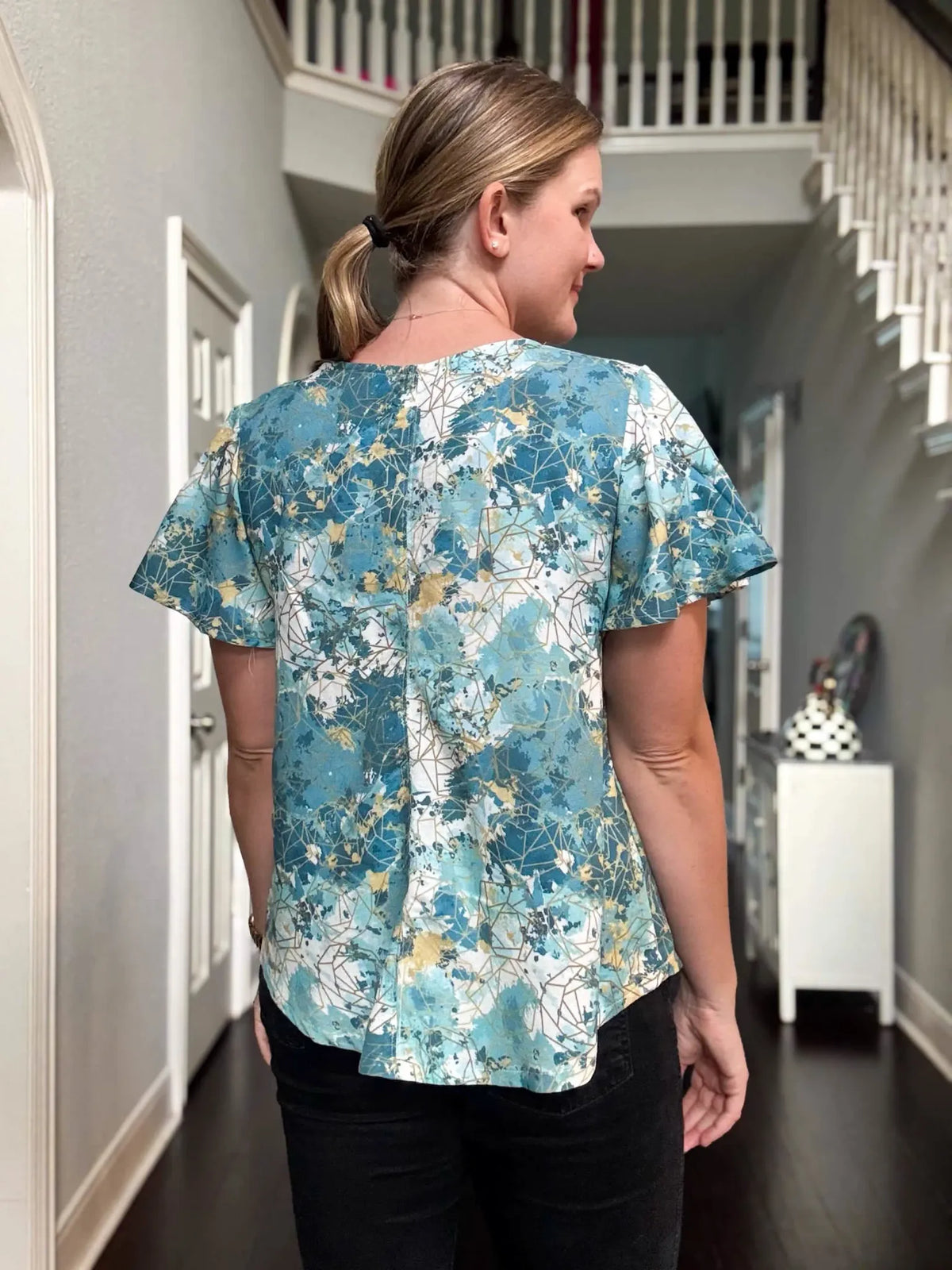 A woman from the back wearing a blue, white, and gold patterned top and black pants.
