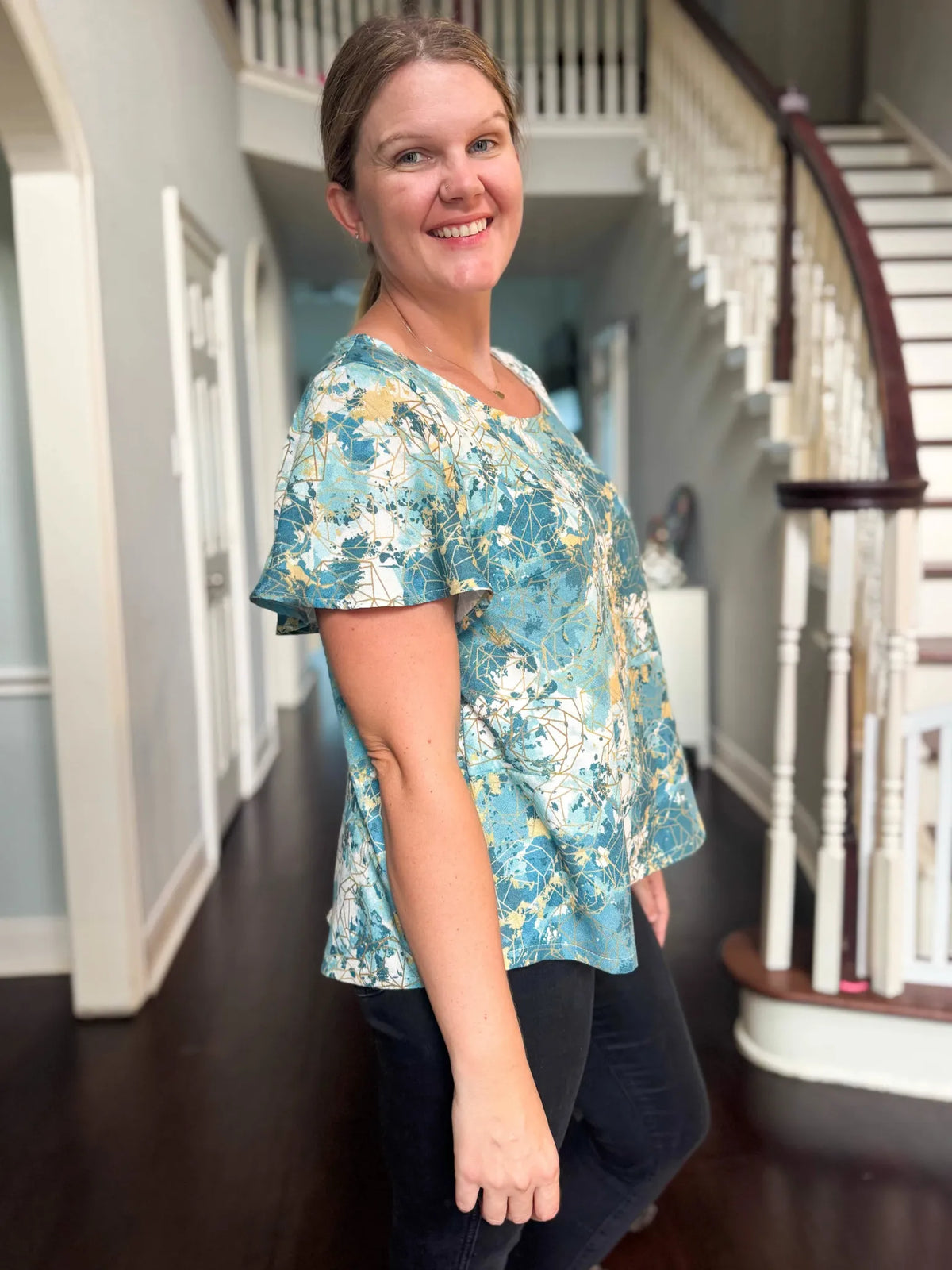 A woman smiles, wearing a blue, white, and gold patterned top and black pants.