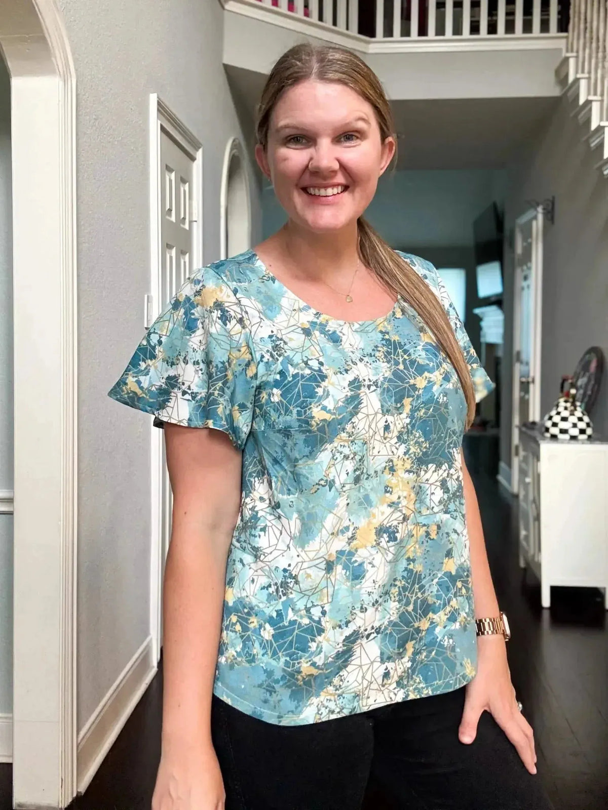 Woman wearing a teal and gold abstract print blouse with bell sleeves in a home setting.