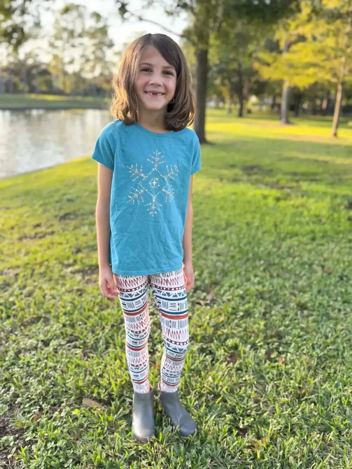 Girl wearing blue snowflake t-shirt, patterned leggings, and gray boots outdoors.
