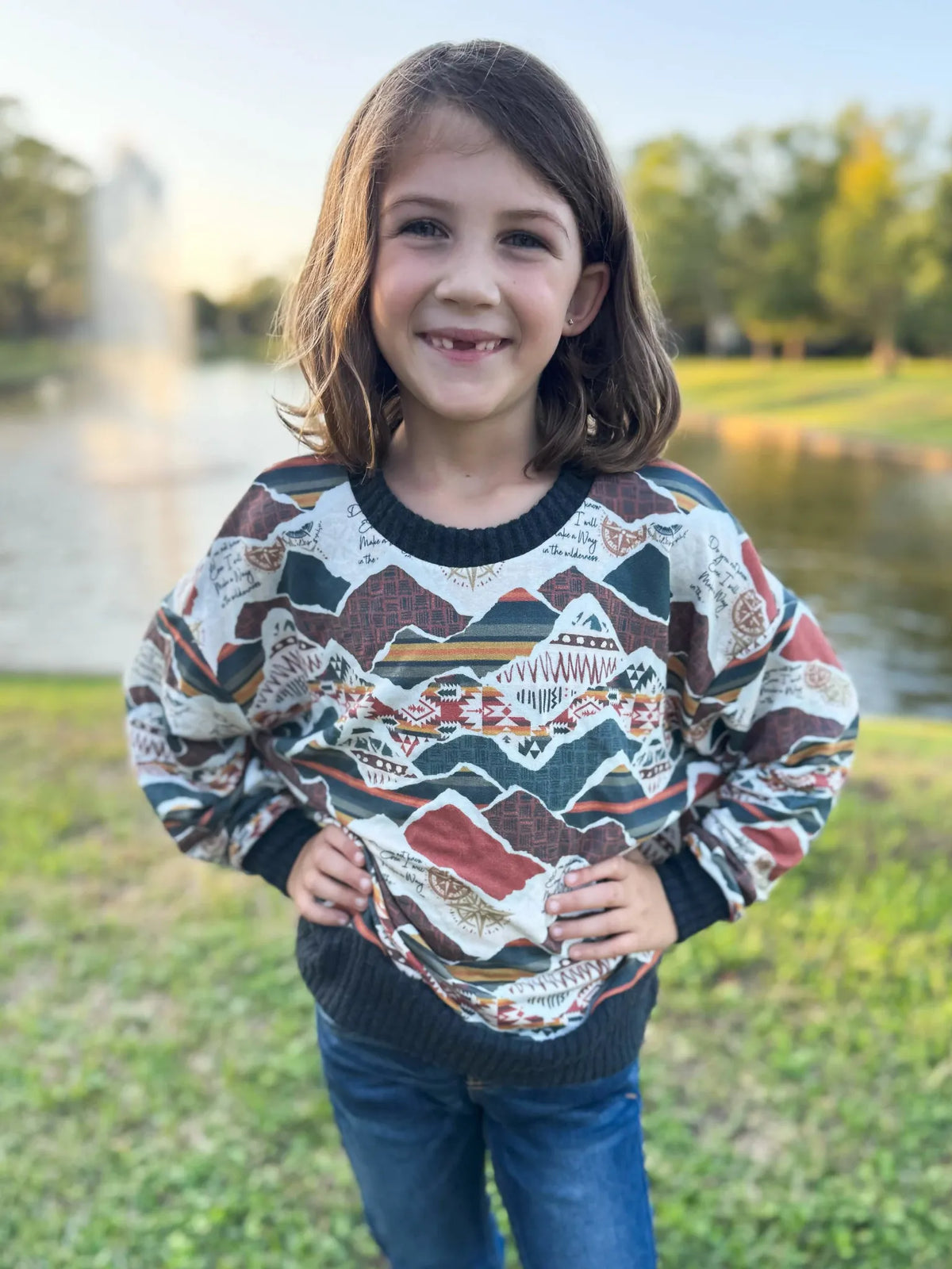 A young girl smiles, wearing a long-sleeved shirt with a mountain and compass pattern.