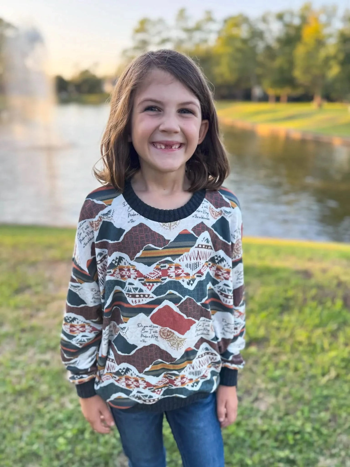 A young girl smiles, wearing a long-sleeved shirt with a mountain and tribal pattern.