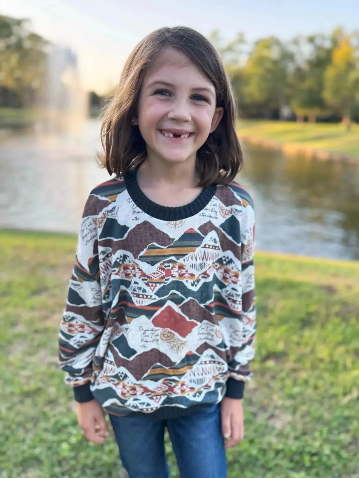 Girl wearing a long-sleeved sweater with multicolored mountain print by a lake.
