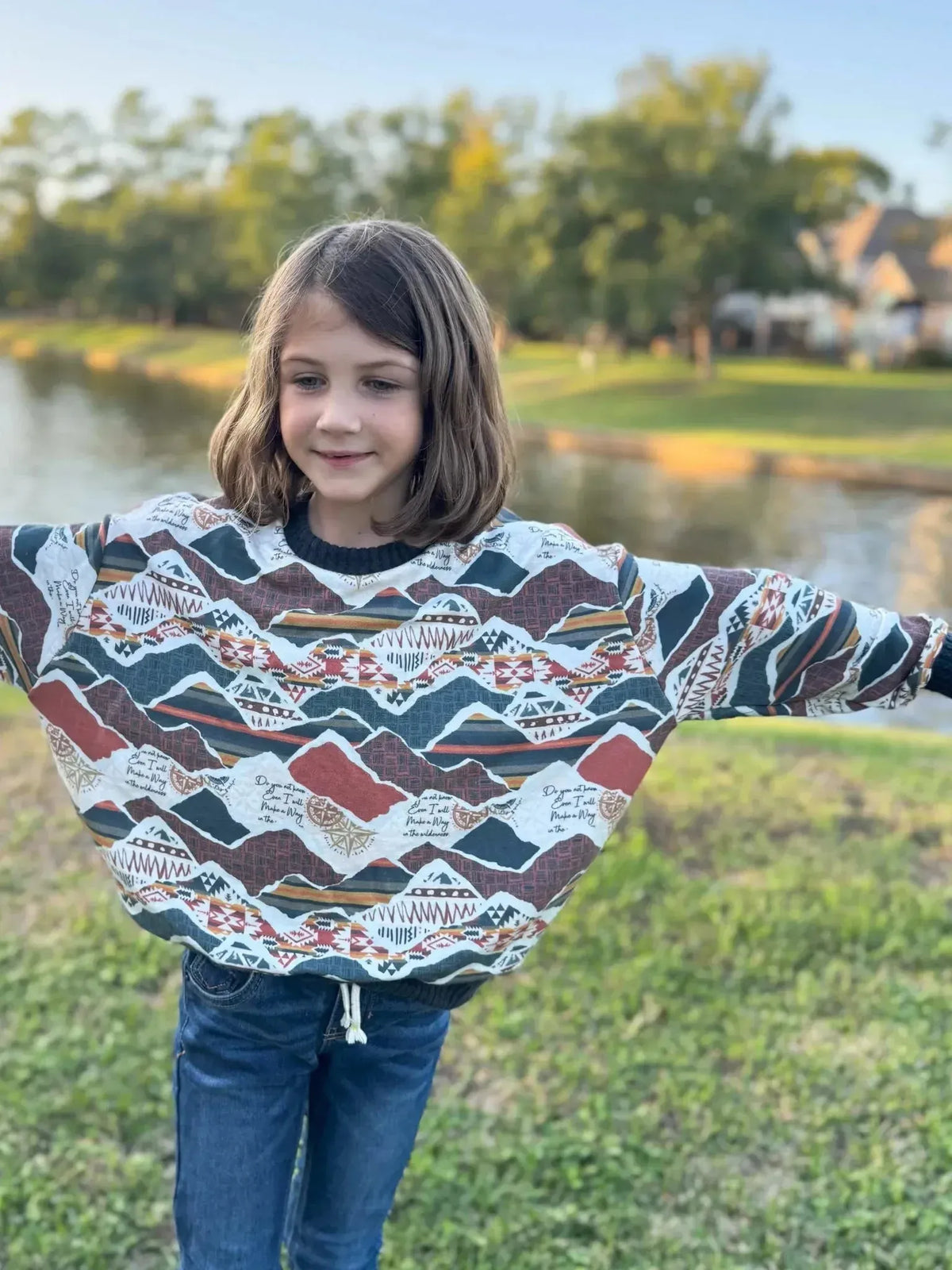 Girl wearing oversized sweatshirt with multicolored mountain print by lake.