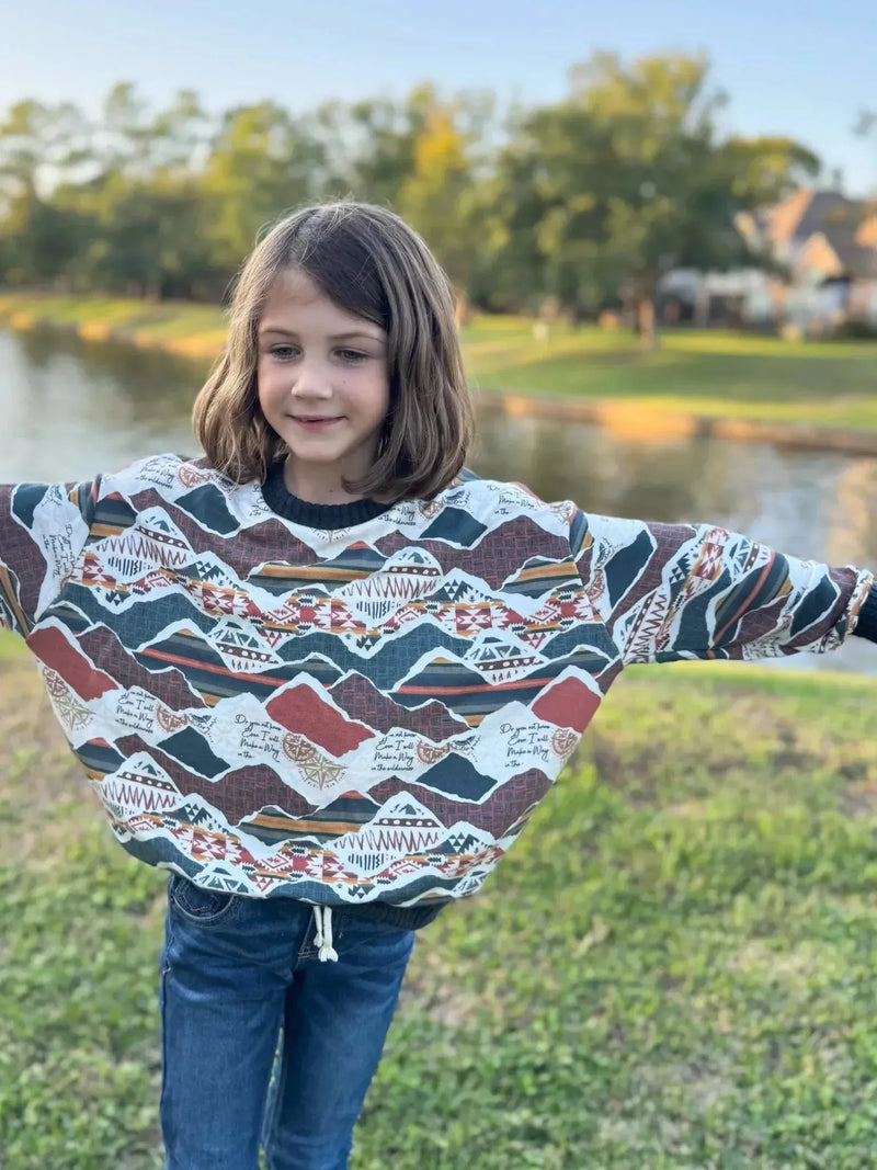 Girl wearing oversized sweatshirt with multicolored mountain print by lake.